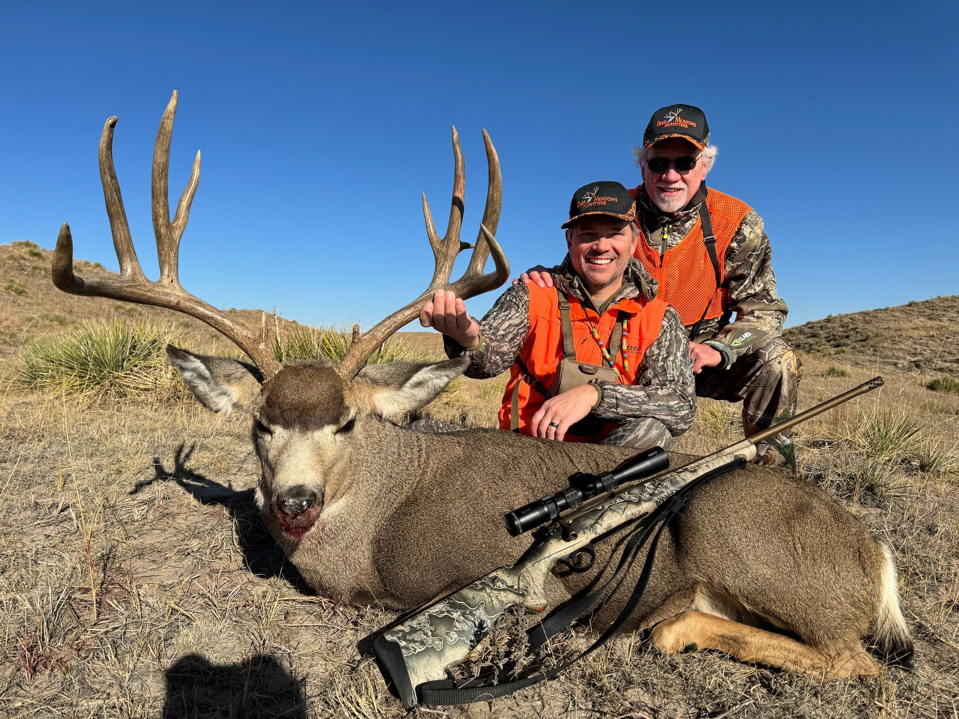 Two men pose with a large deer they hunted, outdoors. One kneels, the other stands, both wearing orange vests.
