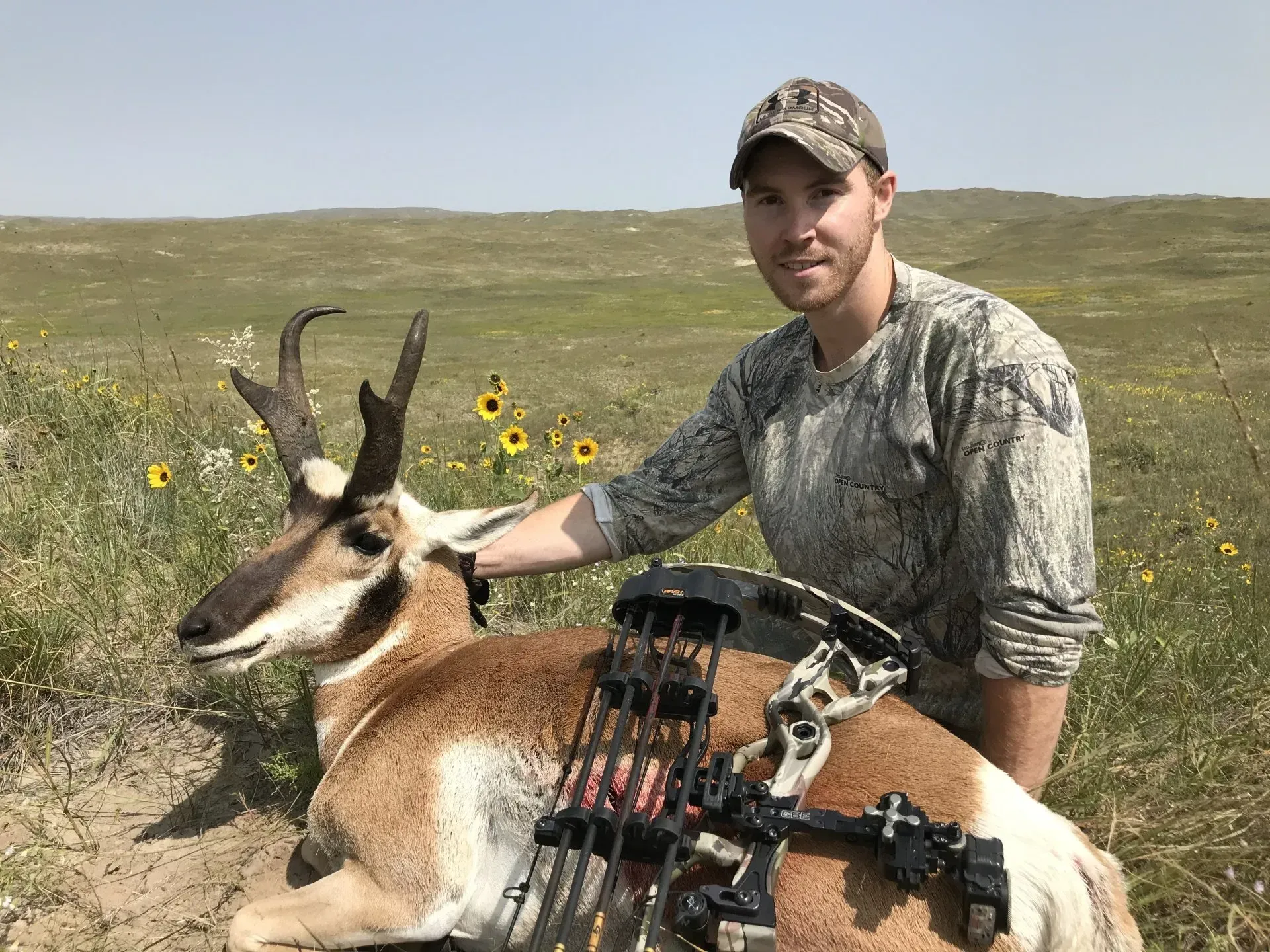 Man in camouflage with bow next to a pronghorn on a grassy plain.