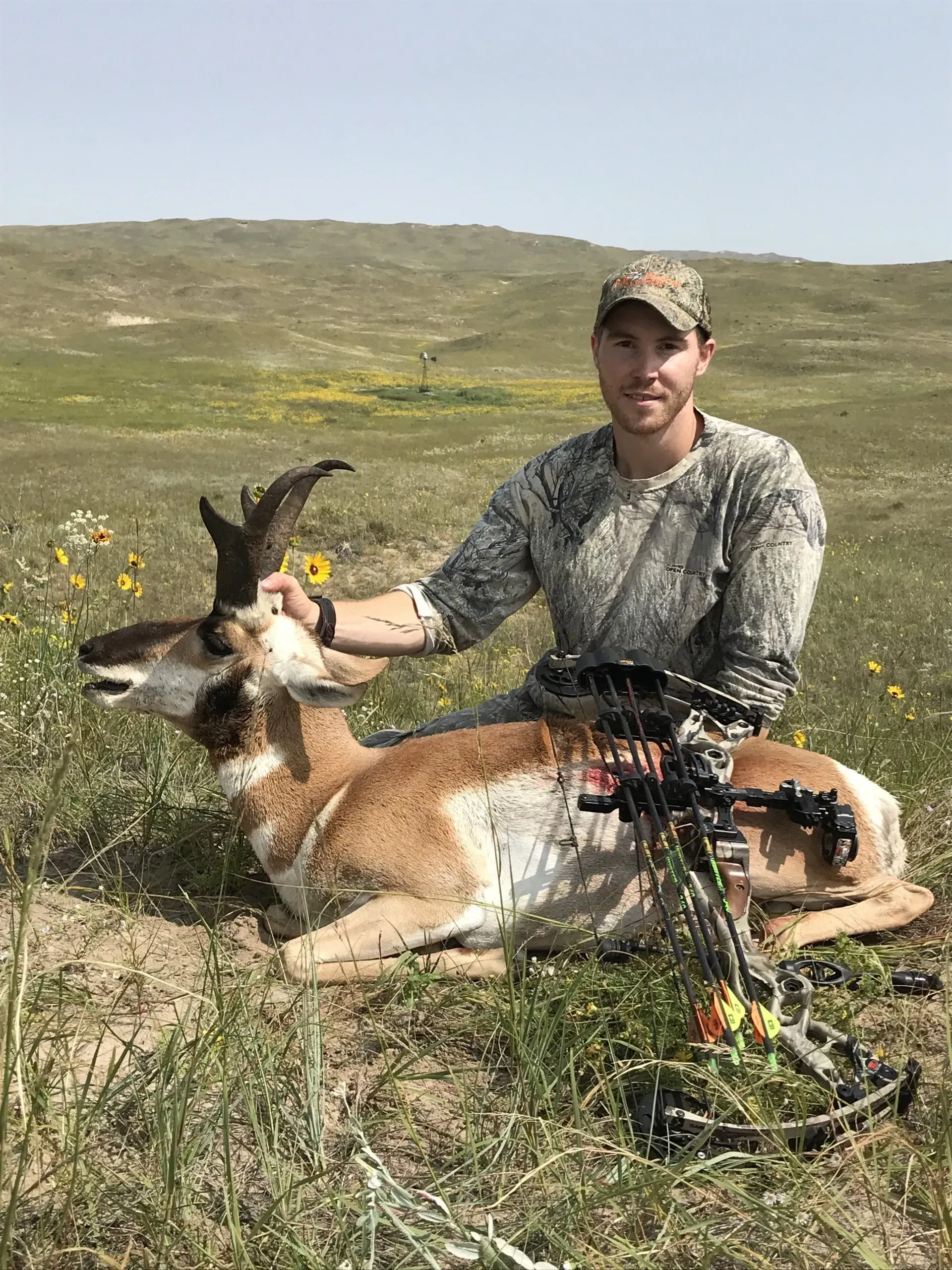 Man in camo with pronghorn antelope on grassy plains, bow in foreground.