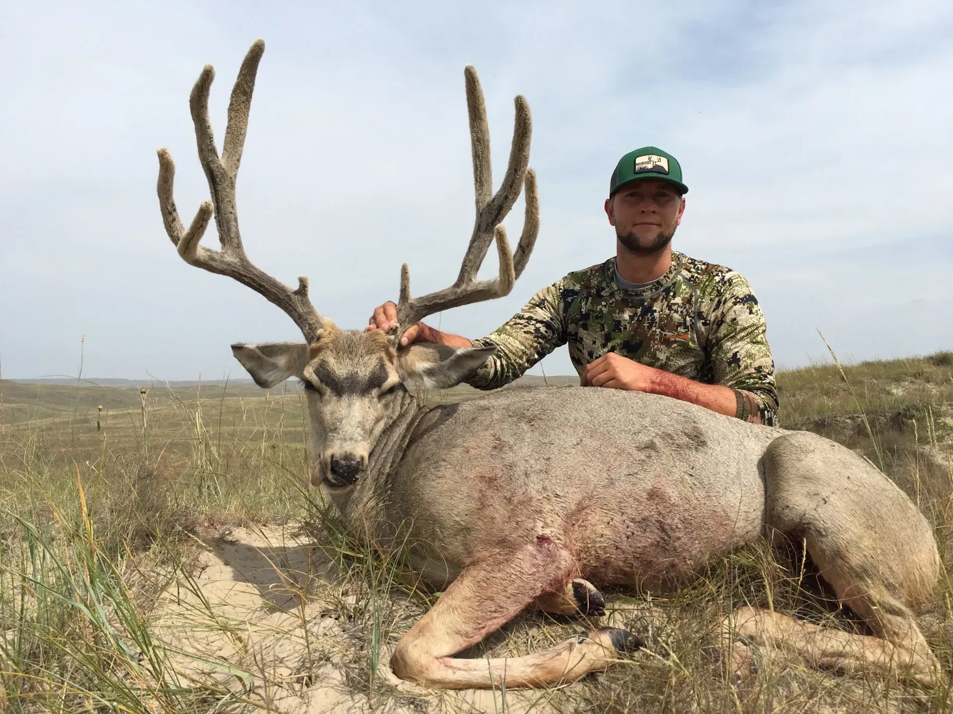 Man in camo with a large buck, sitting in tall grass. The buck has huge antlers; the setting is outdoors.