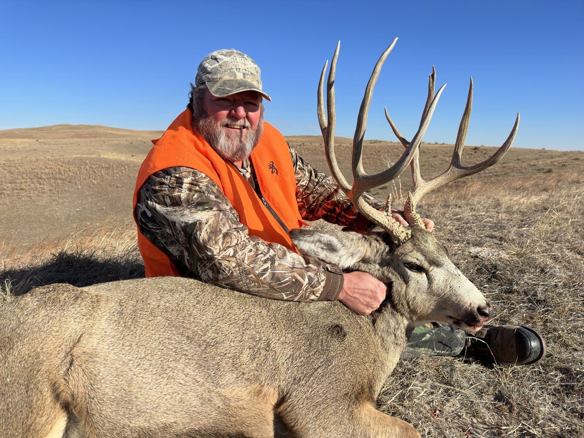 Man in orange vest smiles, holding a large buck with impressive antlers in a field.