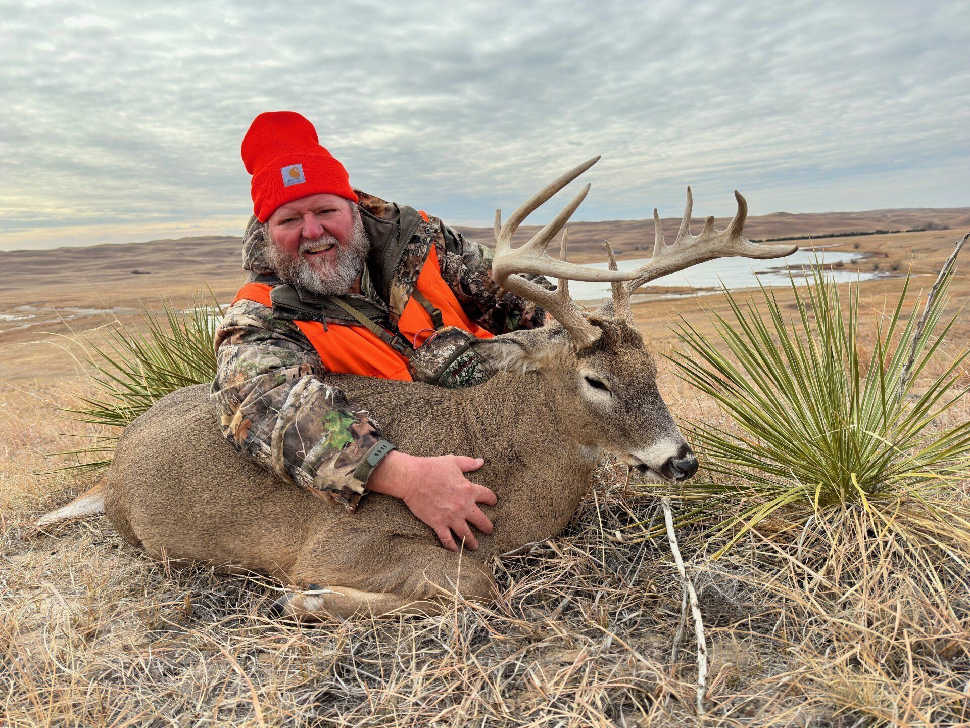 Man in hunting gear smiles, hugging a large buck. They are in a grassy field.