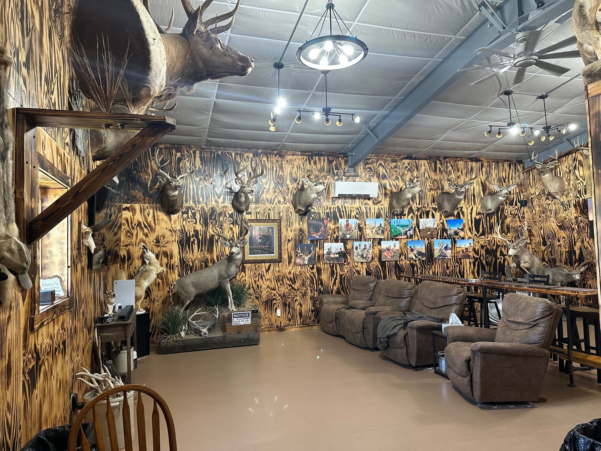 A room with taxidermied animals on walls, a deer head mounted, brown recliner chairs, and a bar table.