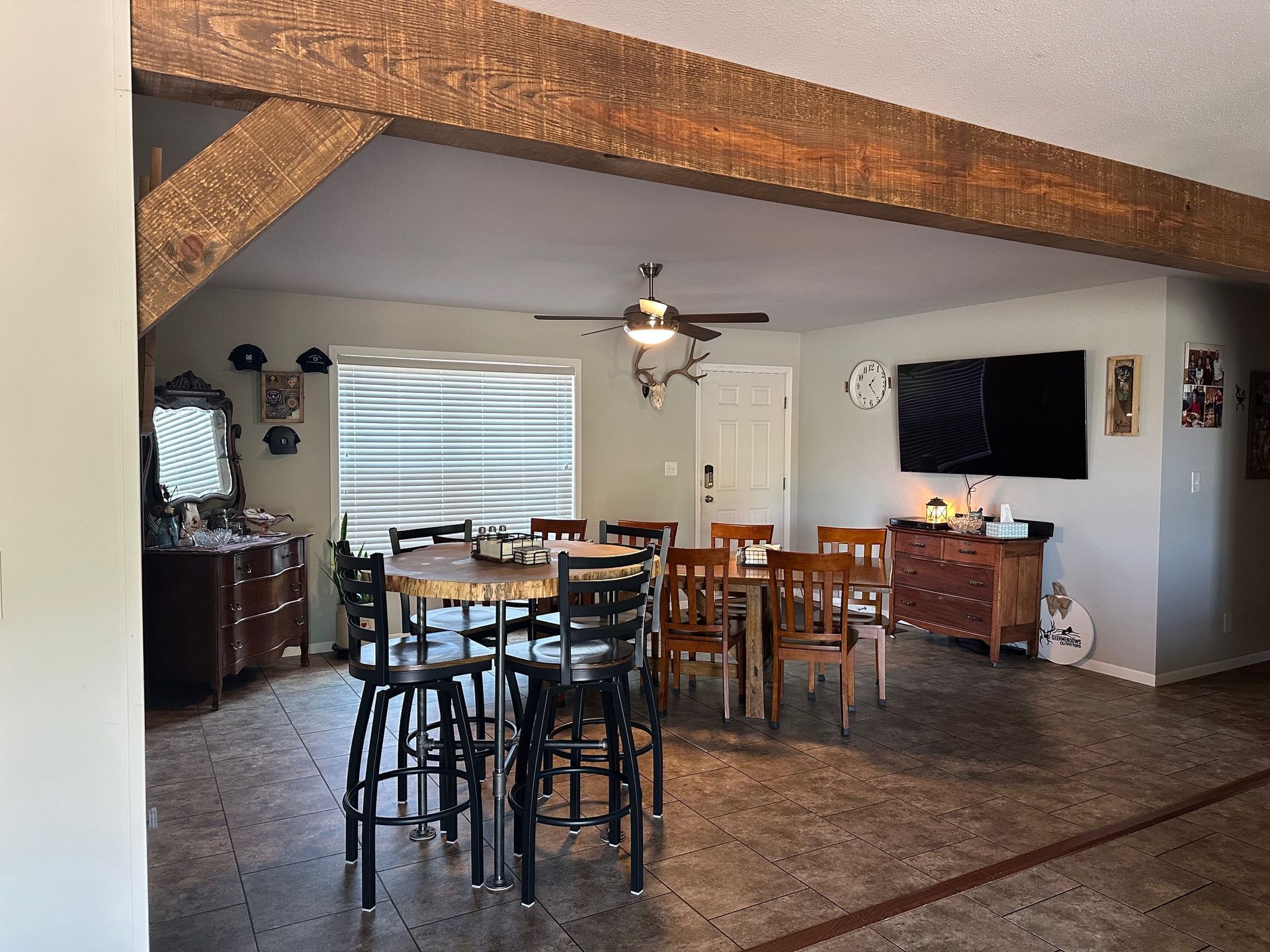 A dining area with a round table, chairs, and a TV on the wall.  Dark brown wooden beams overhead.