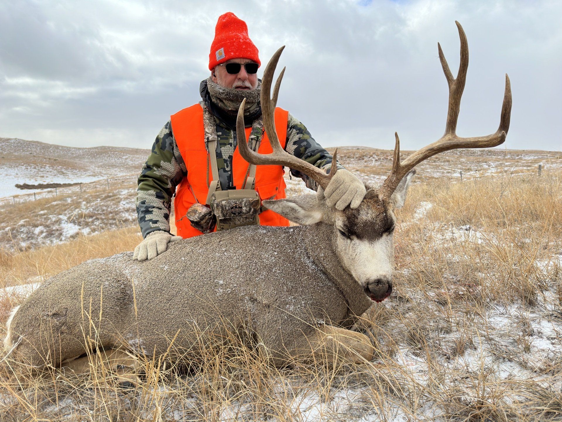 Hunter in orange vest, with large buck. Cold, snowy landscape. Man with sunglasses, orange hat smiles.