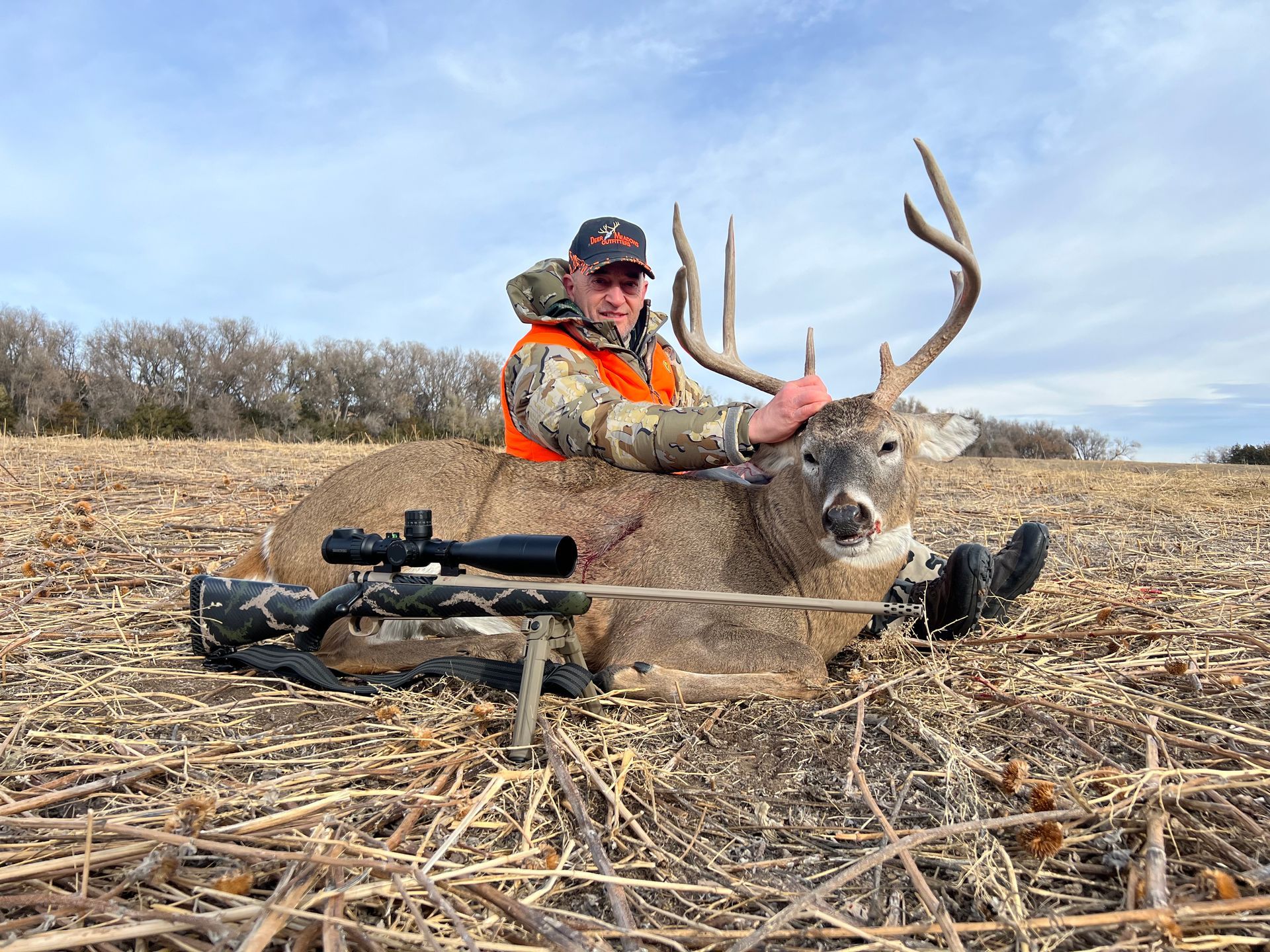 Hunter in orange vest and camo jacket with large buck he harvested on a field. Rifle with scope is visible.