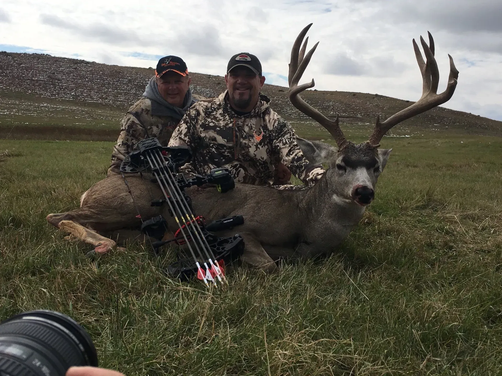 Two men pose with a large buck deer in a grassy field; they are in camouflage, a bow and camera are in the foreground.