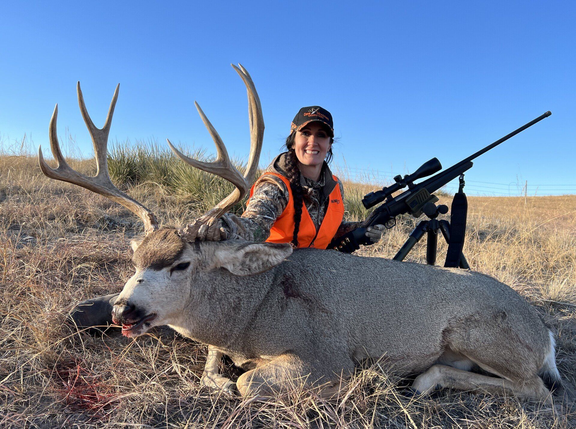 Woman in orange vest with rifle, poses with a large buck in field on a sunny day.