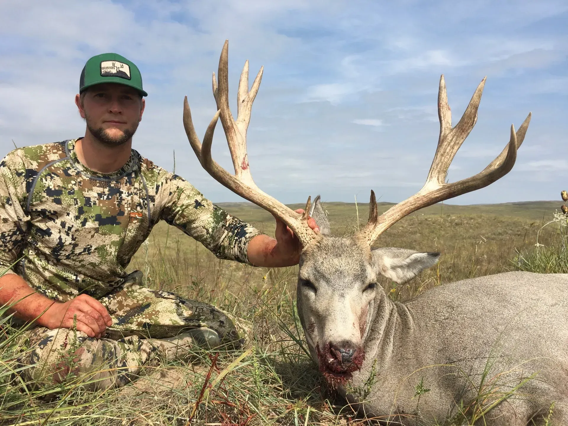 Man in camo with a large buck he harvested. The buck has large antlers, is on the ground in a field.