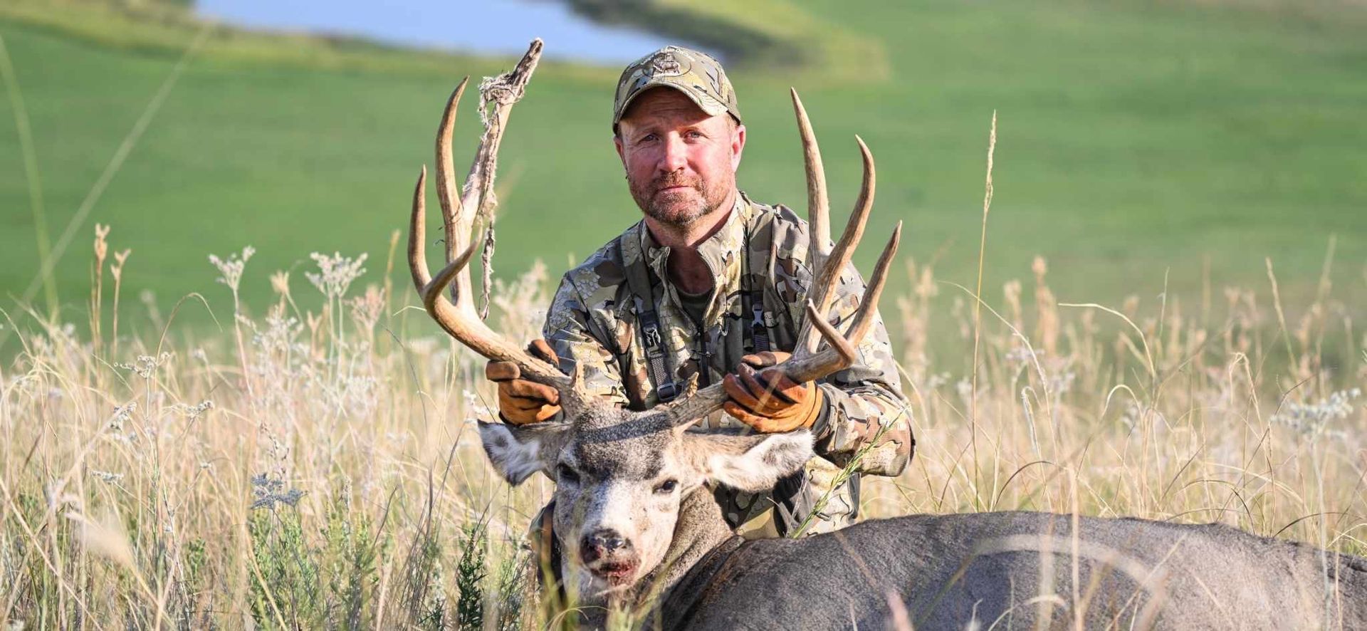 Man in camouflage holds a large buck with impressive antlers in a field, smiling at the camera.