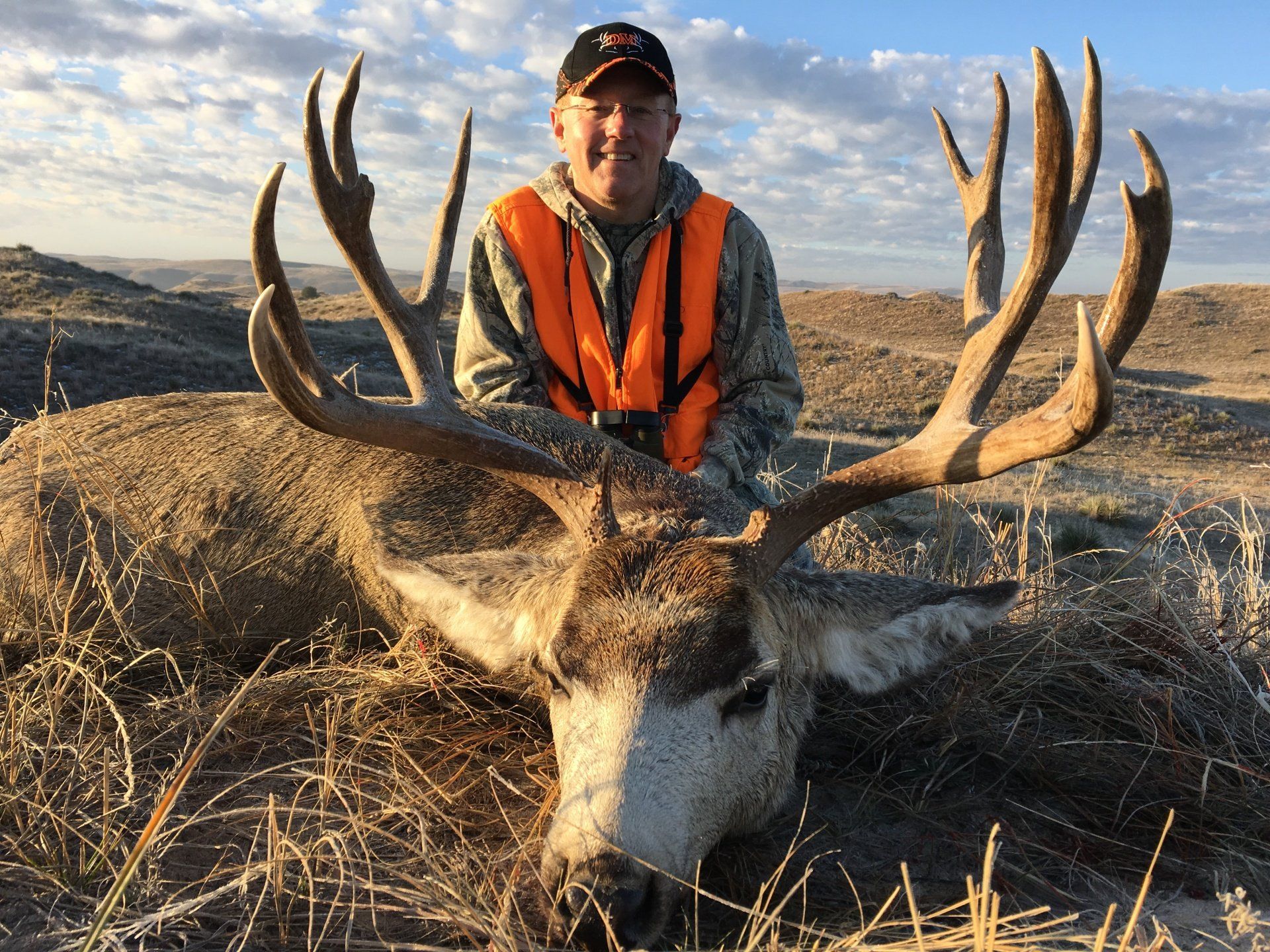 Man in orange vest poses with a harvested deer, large antlers, in a dry field.