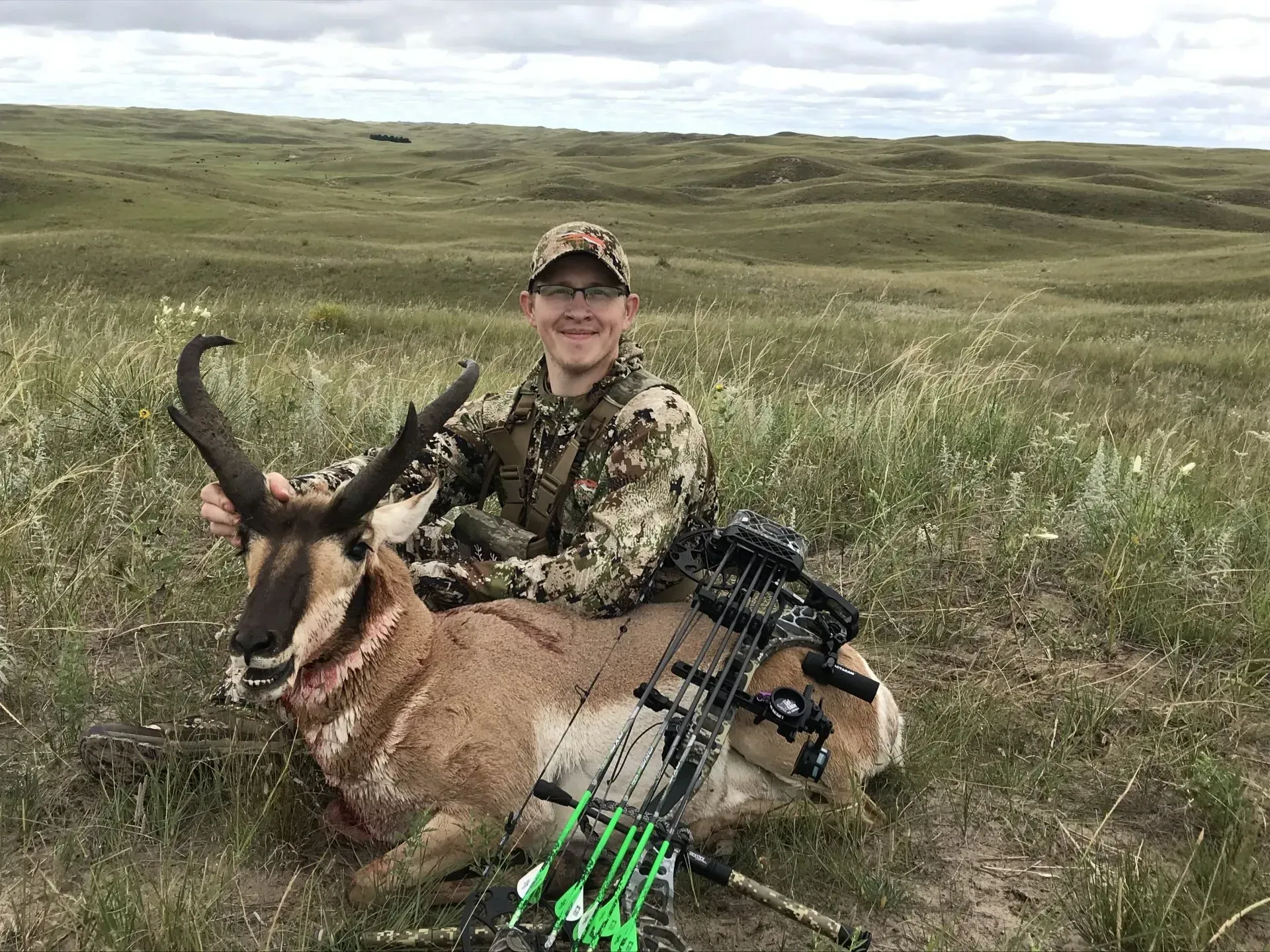 Man in camo holds a pronghorn antelope he hunted in a grassy field. A bow lies beside it.