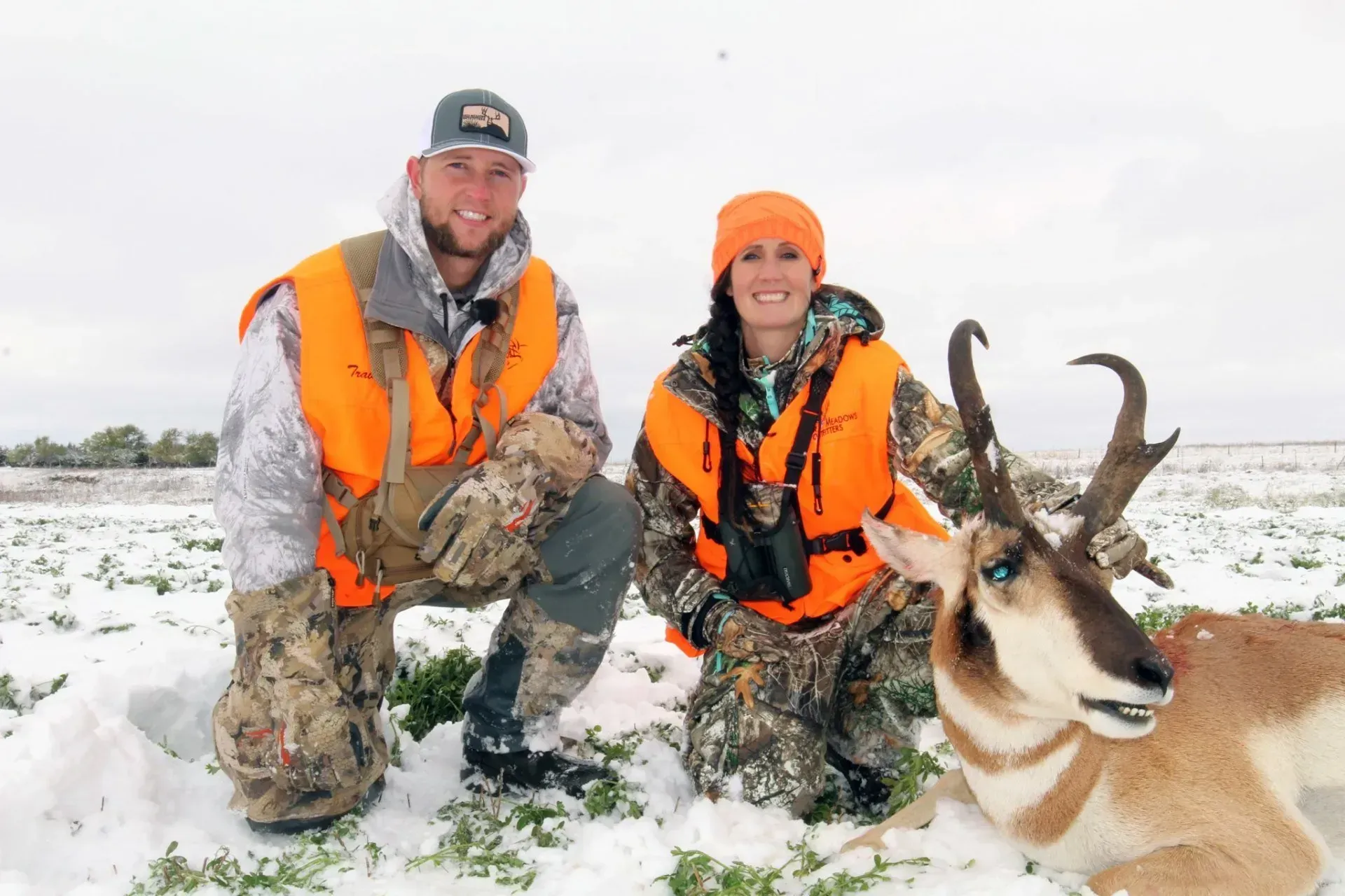 Two hunters kneel beside a pronghorn antelope in a snowy field. Both wear orange vests.