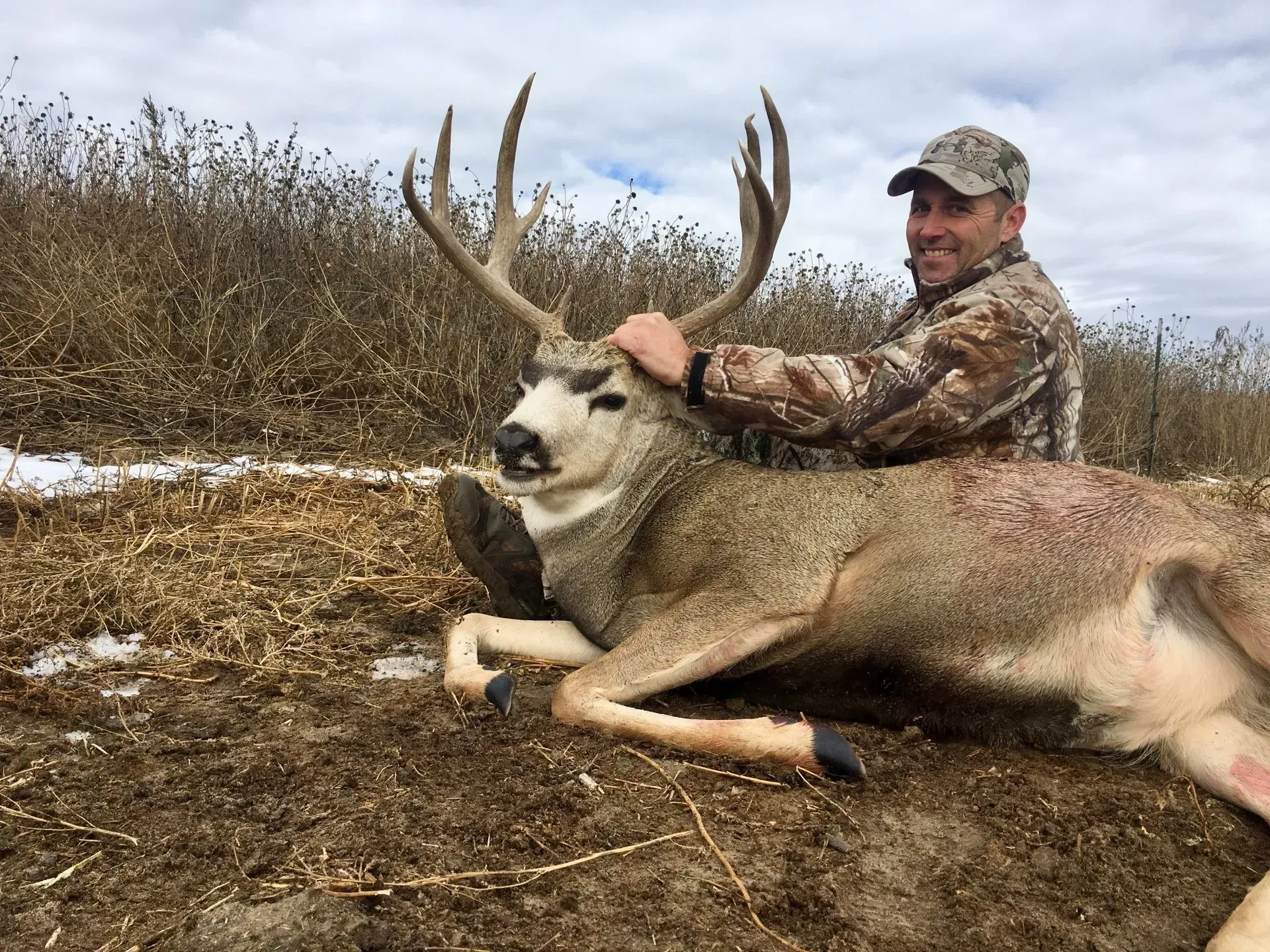 Man in camo smiles, petting a large dead buck with large antlers. Field setting, cloudy sky.