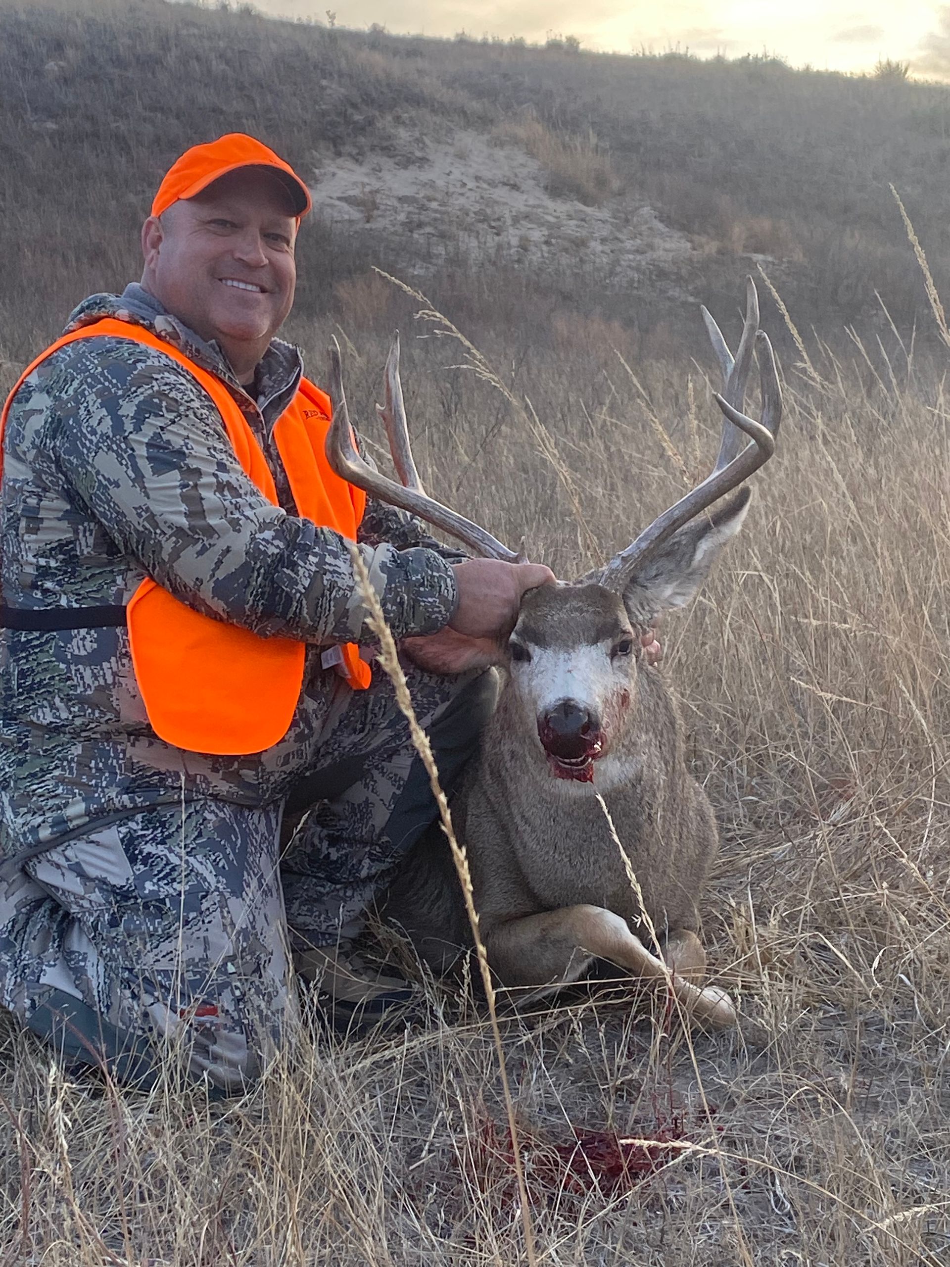 Hunter kneels beside a large buck, smiling. Orange vest, camo clothing. Grassy hillside setting.