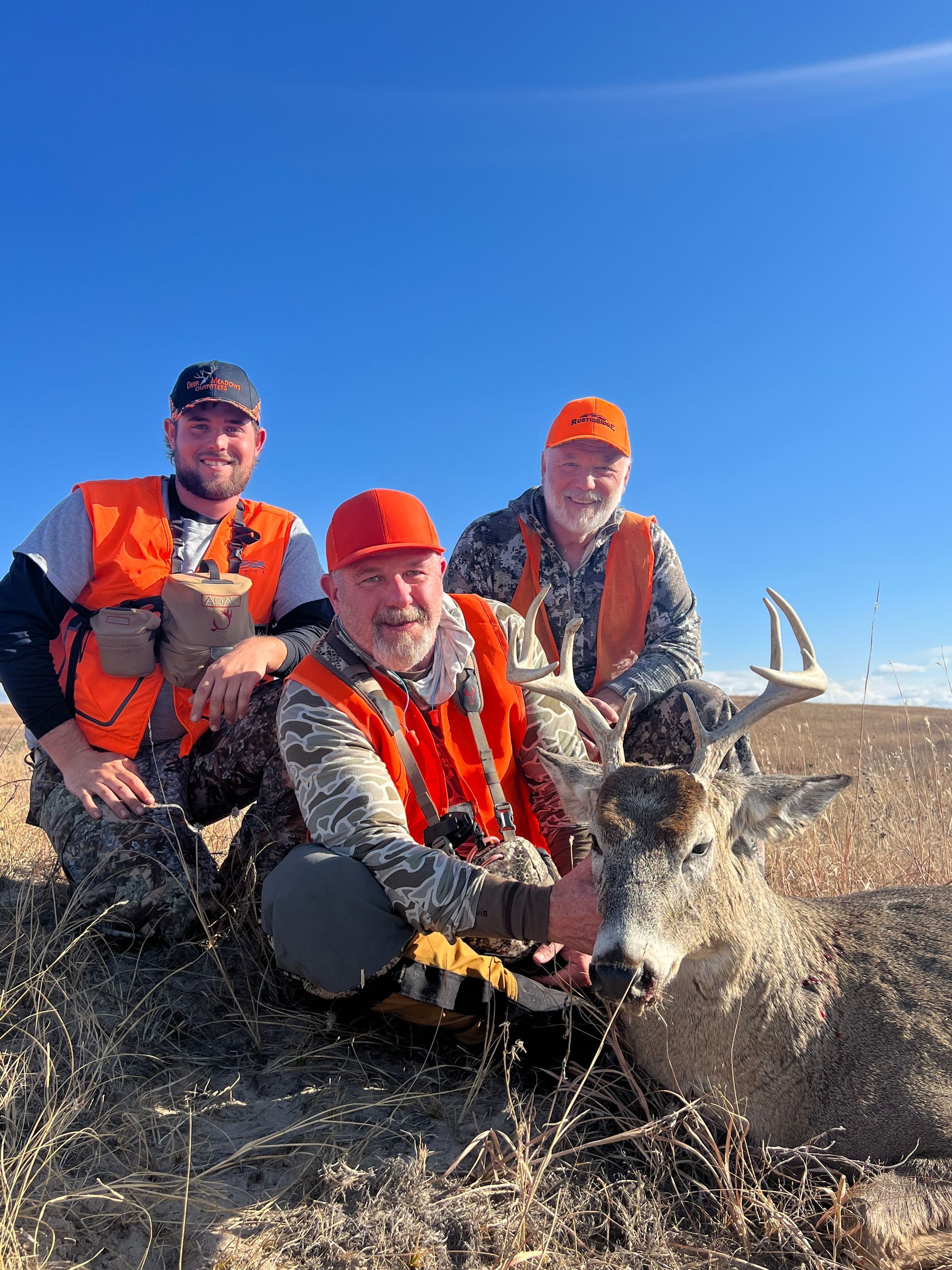 Three hunters in orange vests pose with a harvested deer in a field, under a blue sky.