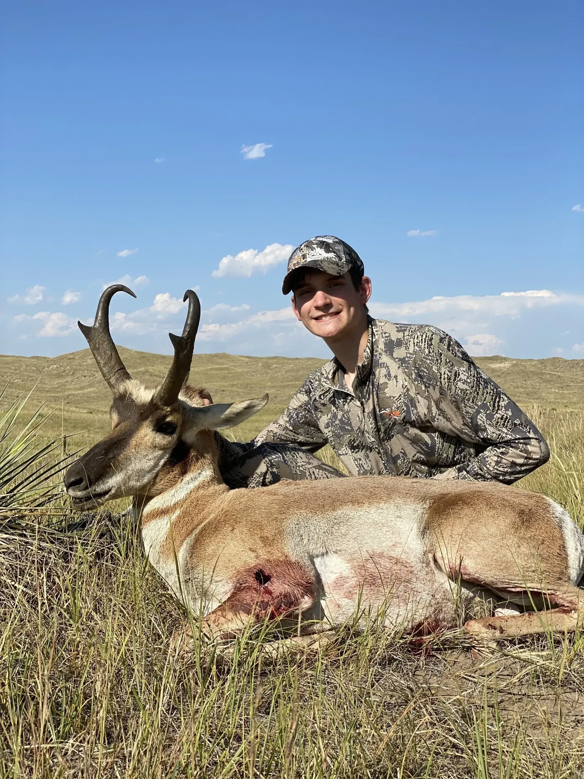 Young hunter smiles next to a harvested pronghorn antelope on a grassy plain, blue sky overhead.