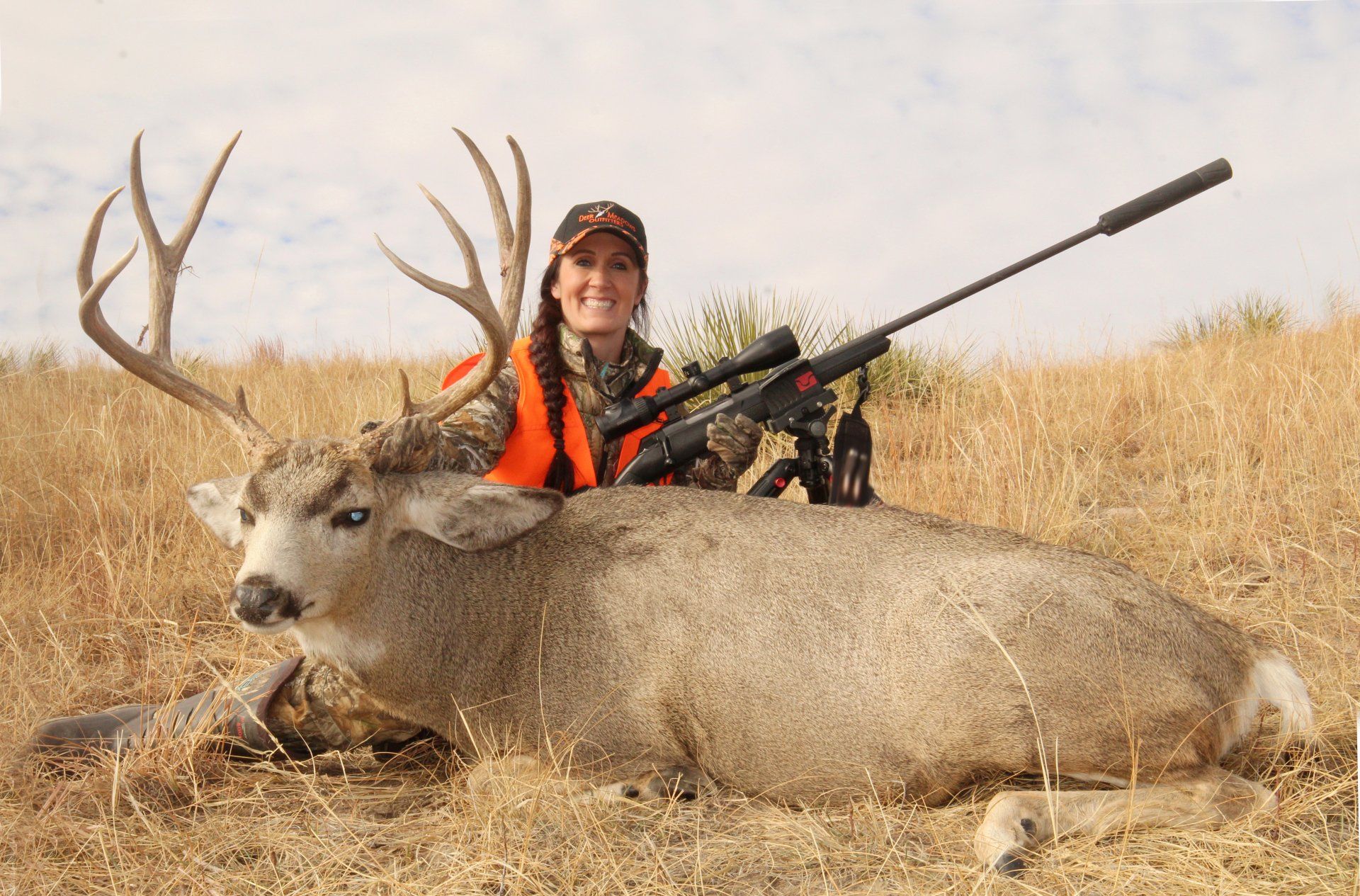 Woman in hunting gear smiles next to a large buck she has shot, rifle in hand, outdoors.