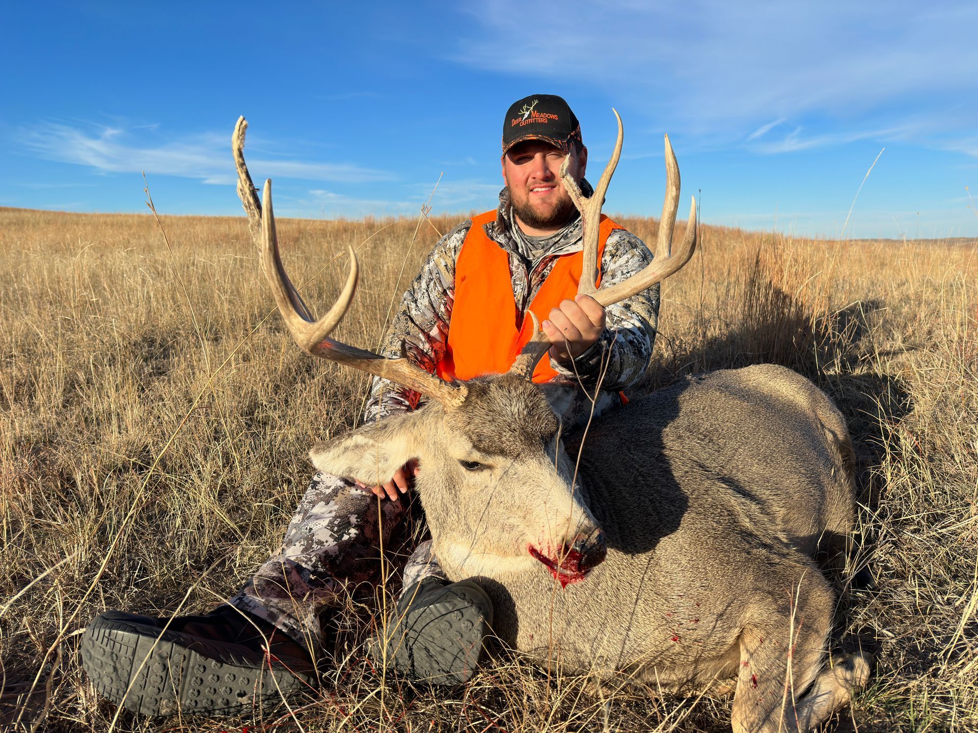Man in orange vest sits with a large buck in a field under a blue sky.