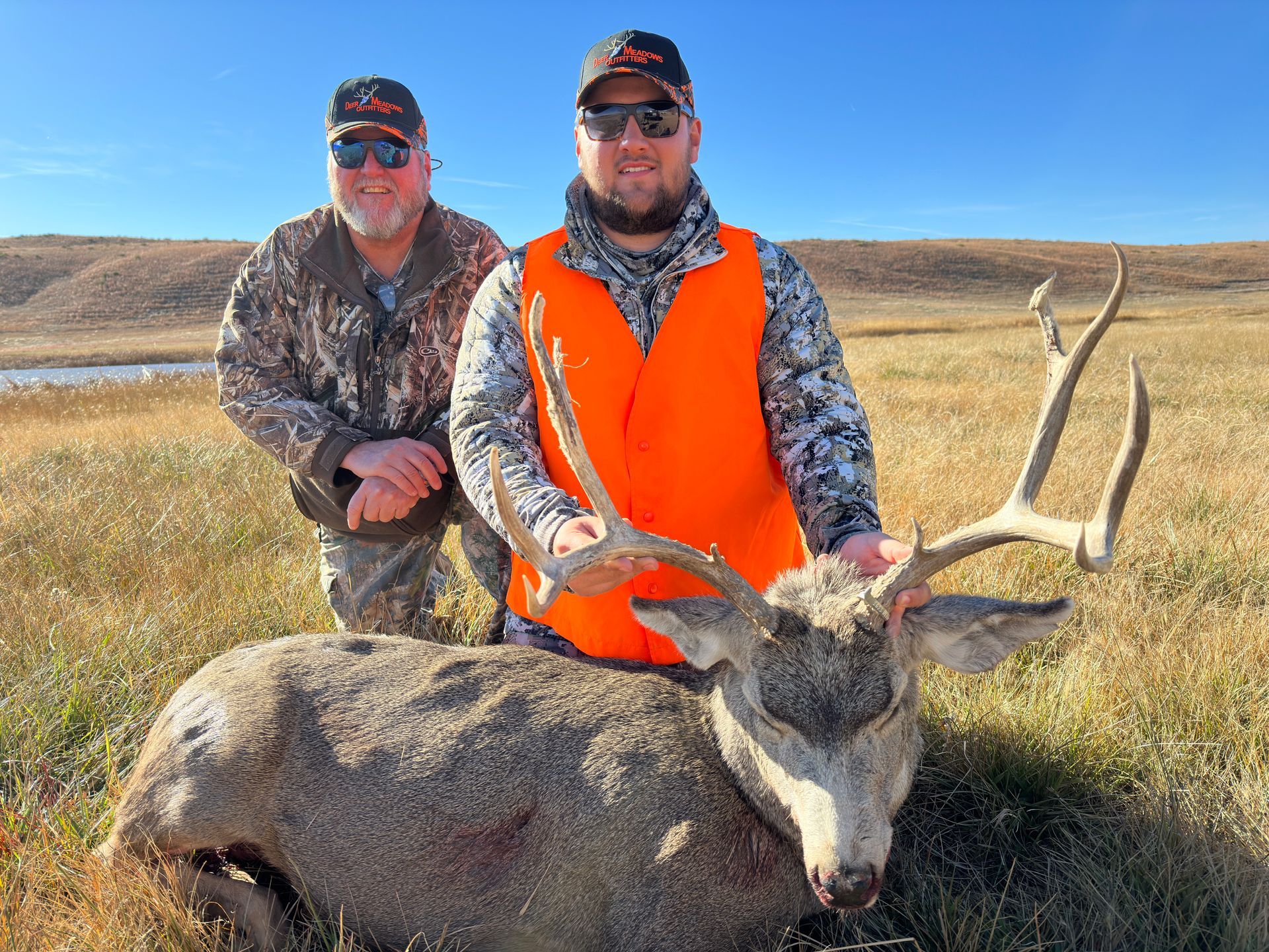 Two men pose with a large buck in a field. One wears orange vest, both wear hats. Sunny day.