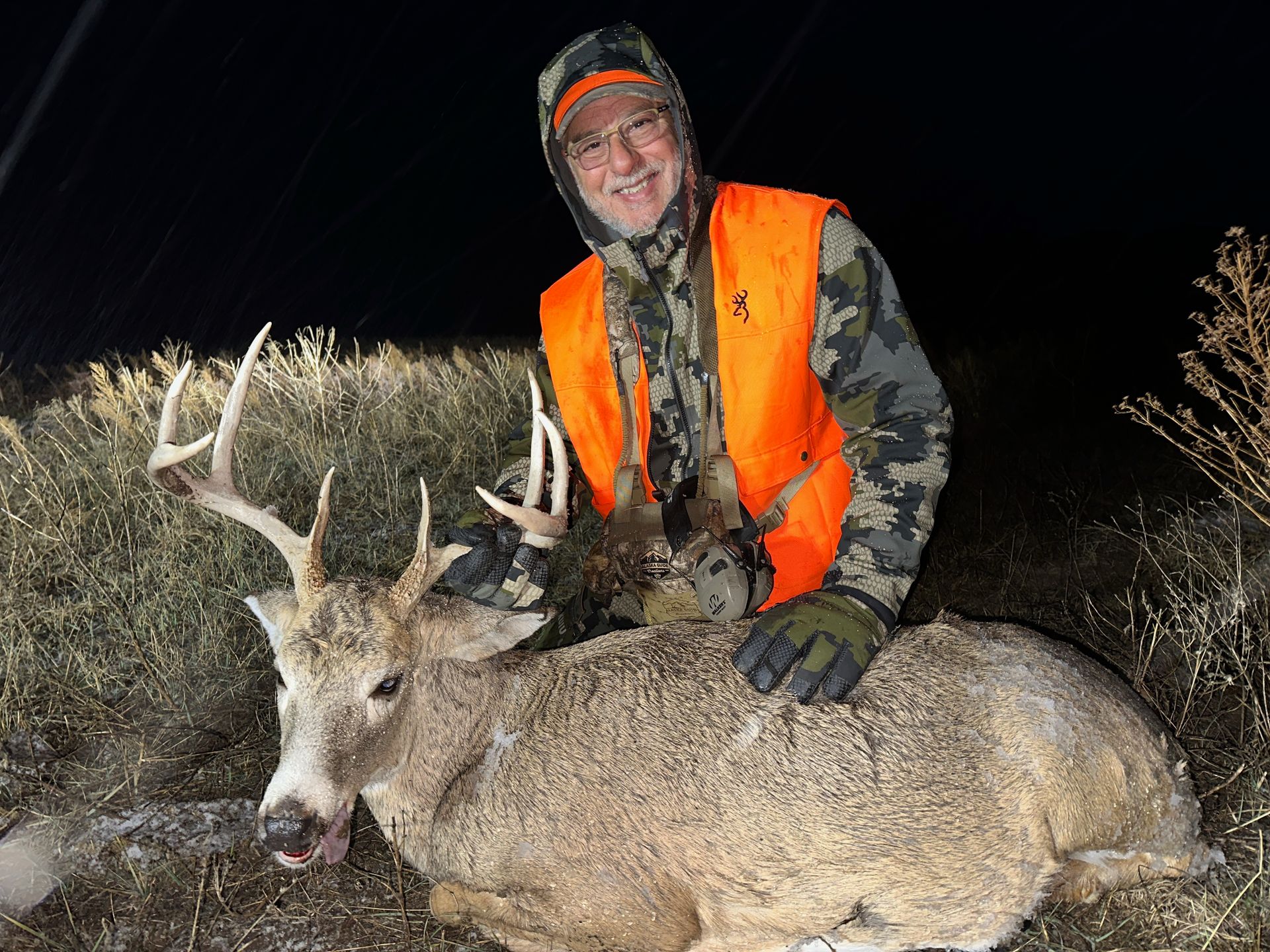 Man in orange vest and camo, kneeling with a large buck he has killed.
