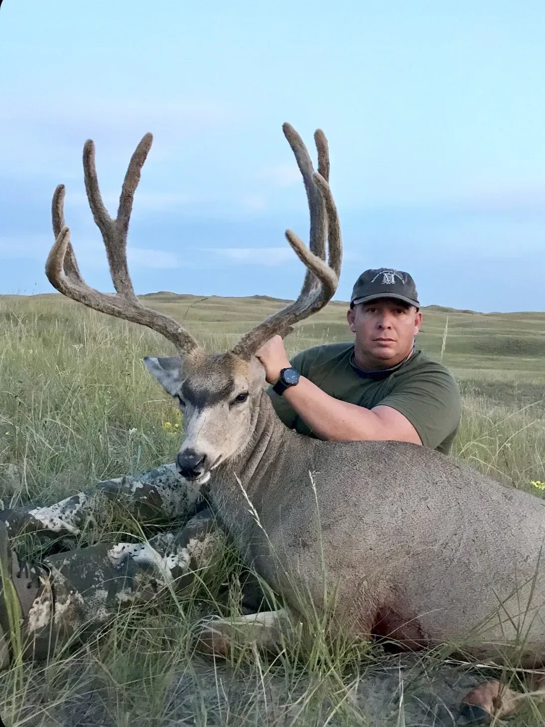 Man in a cap poses with a large mule deer buck he harvested in a grassy field.