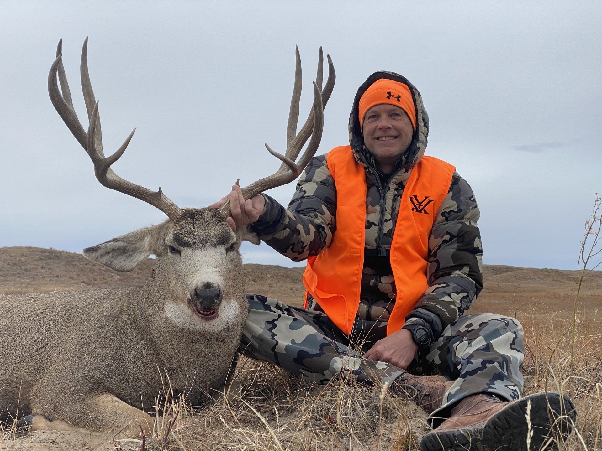 Hunter in orange vest kneels next to large mule deer buck on grassy plain. Cloudy sky.