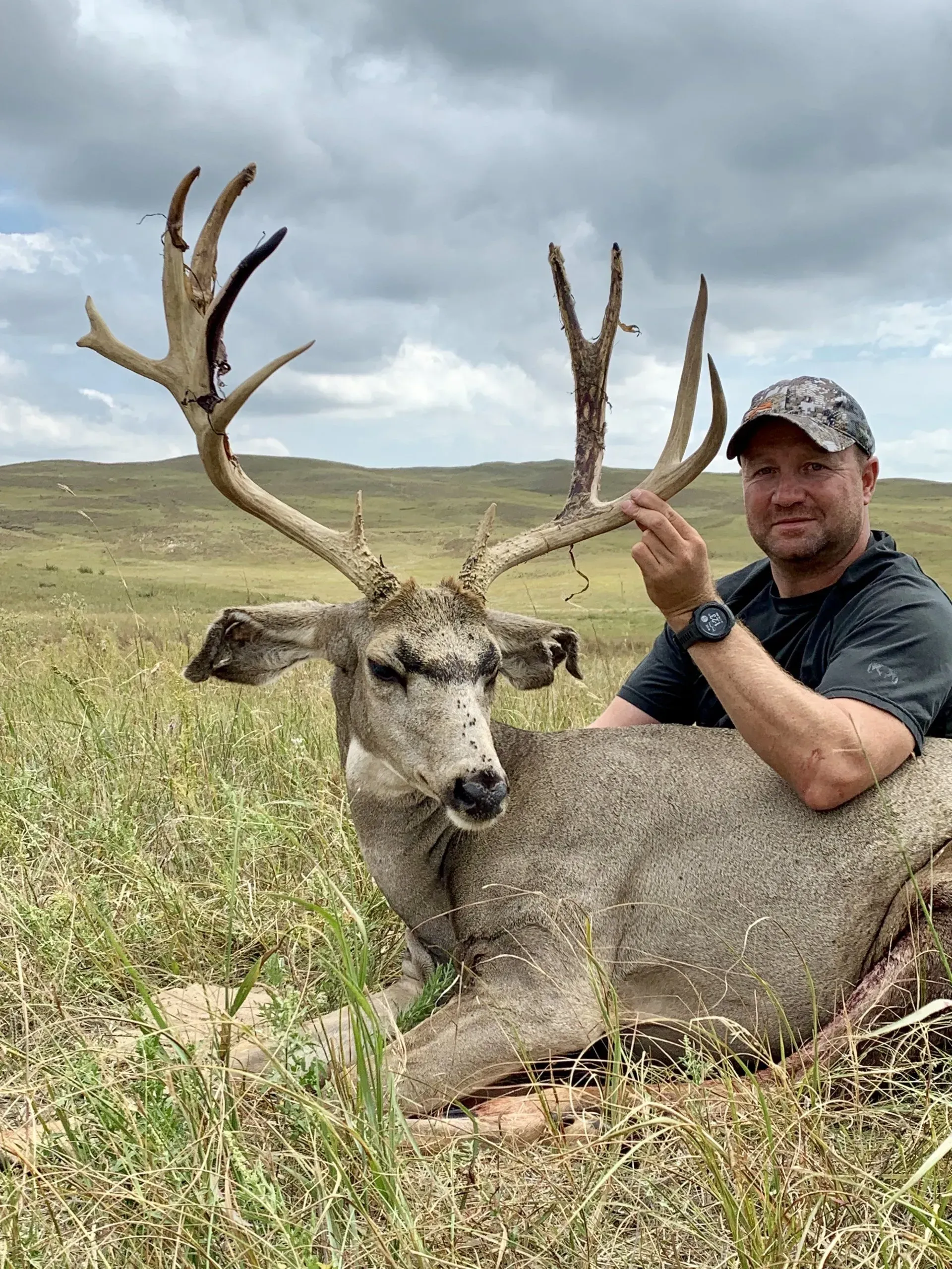 Man poses with a large buck in a grassy field. The man smiles, the deer is lying down, and the sky is cloudy.