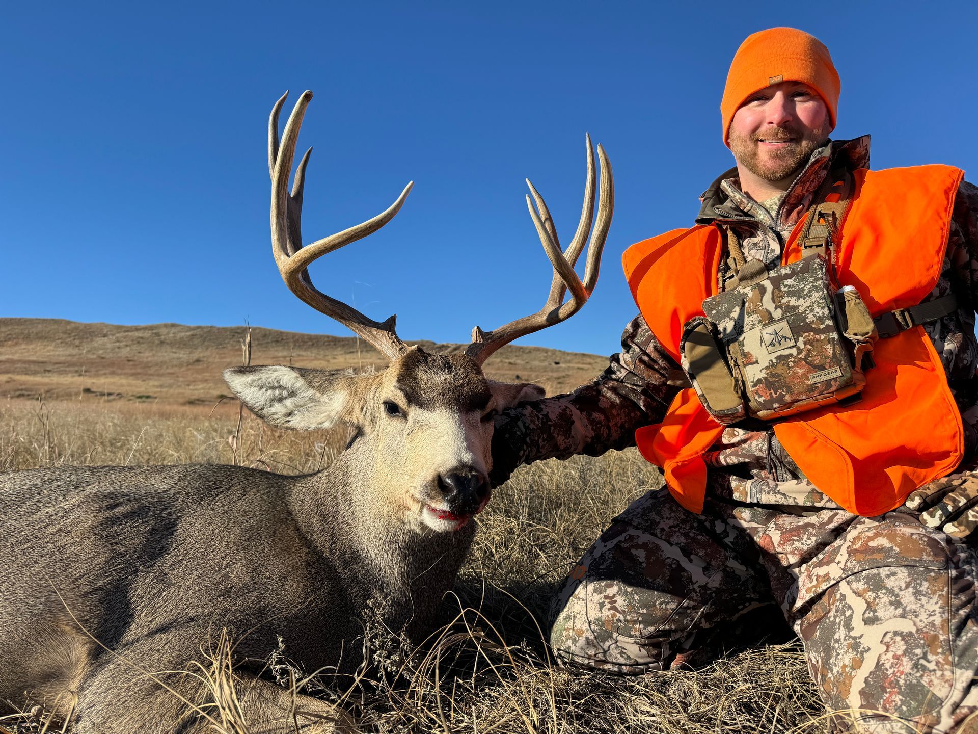 Hunter kneels beside a large mule deer buck with antlers. The hunter wears camo and orange, and smiles outside on a sunny day.