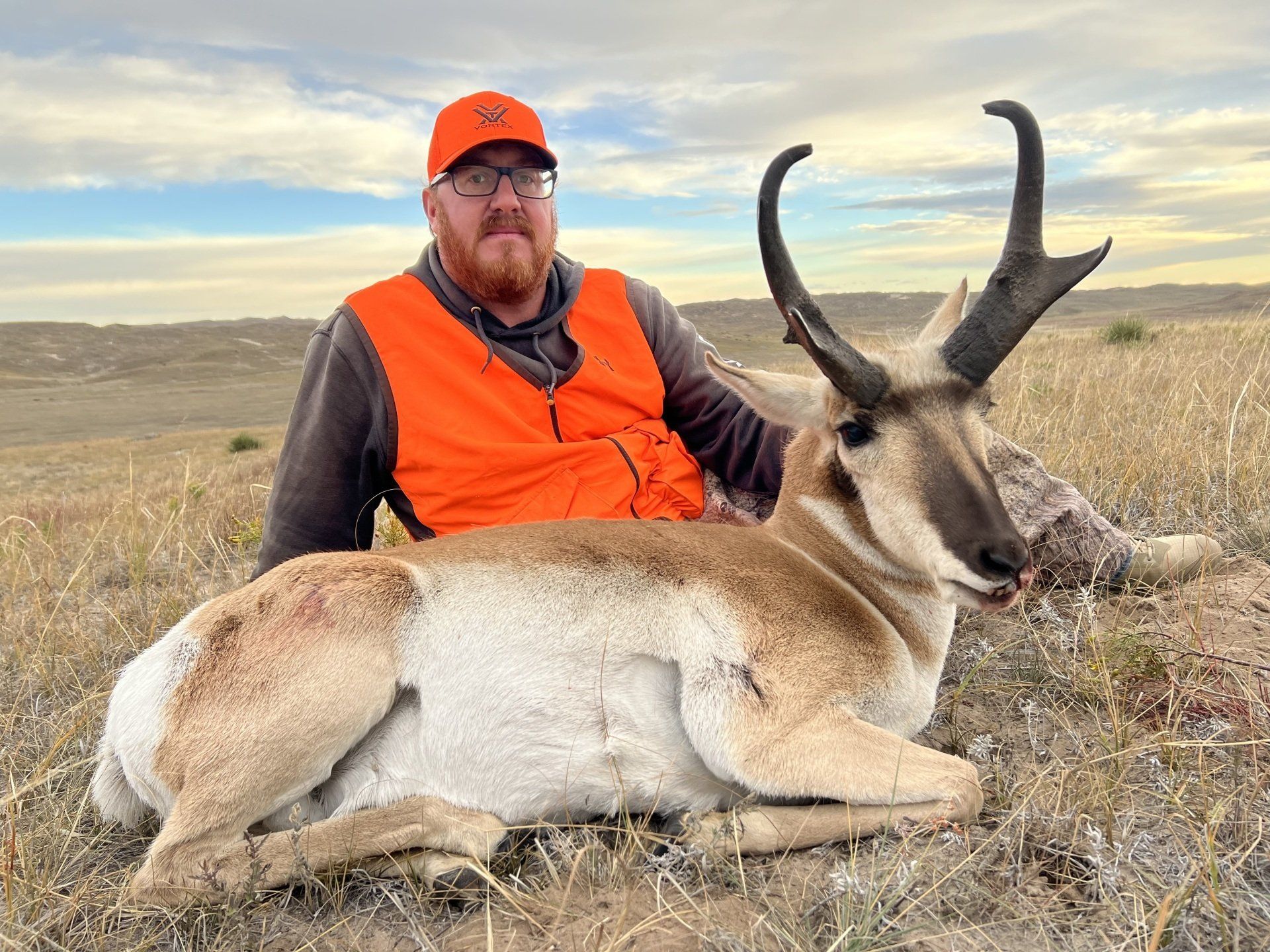 Man in orange vest poses with a pronghorn antelope on grassy terrain.