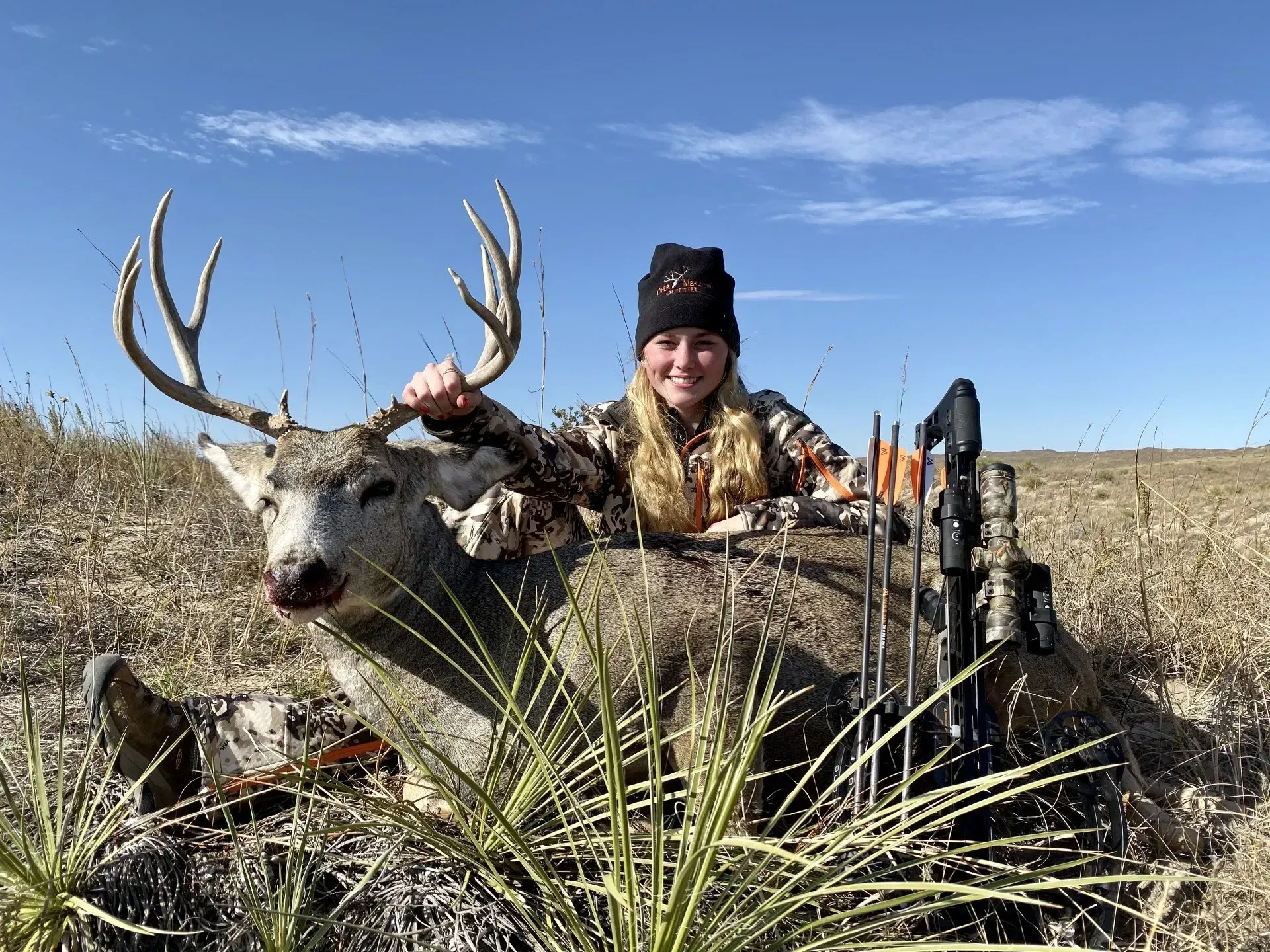 Woman in camo with a large deer, holding its antlers in a field. Blue sky and arrows visible.