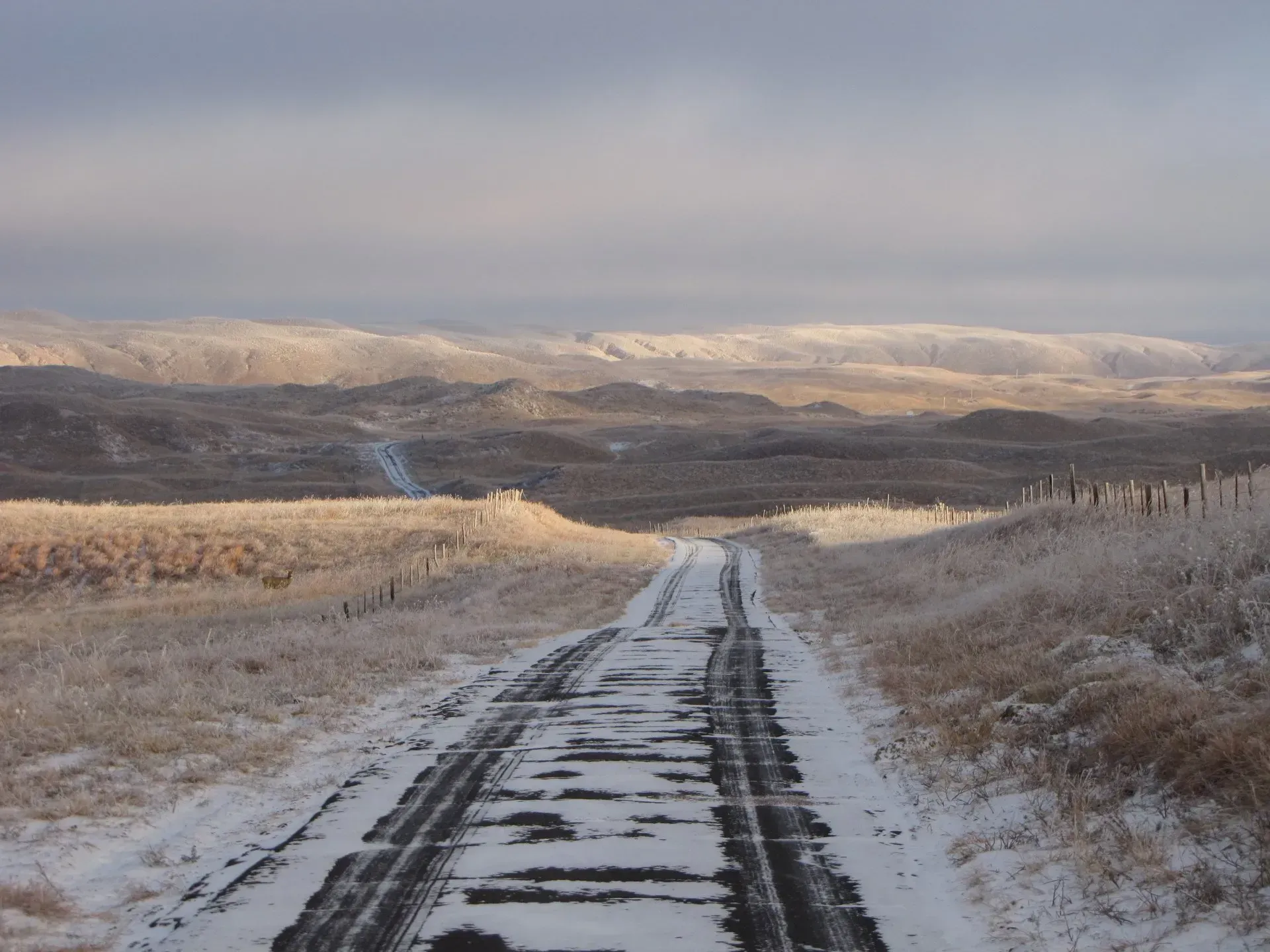 A snow-covered road stretches through a winter landscape toward distant hills under a cloudy sky.