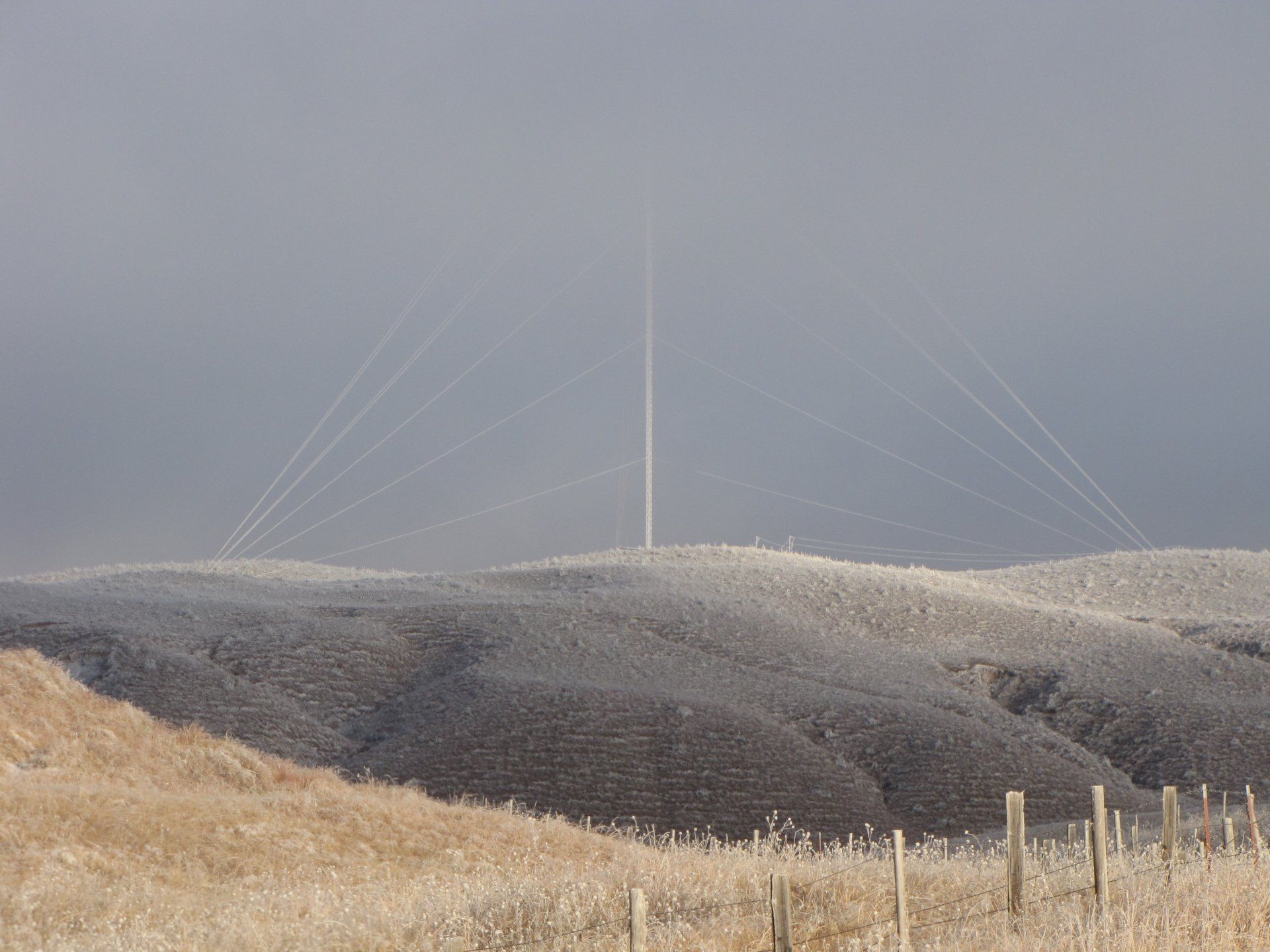 Hilly landscape covered in frost, with radio tower and cables extending into a foggy sky.
