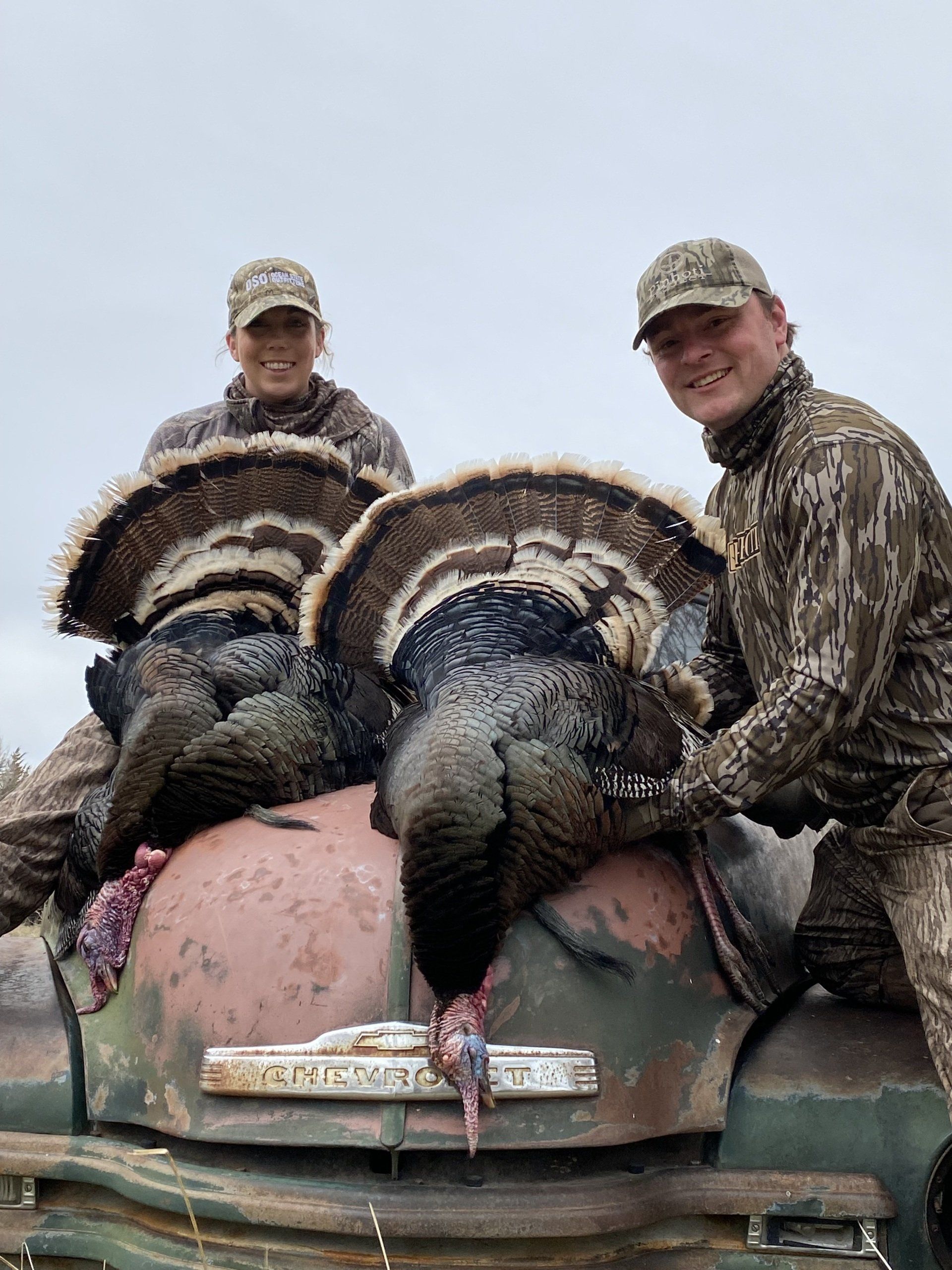 Two hunters, smiling, pose with two turkeys on the hood of an old truck, outdoor setting.
