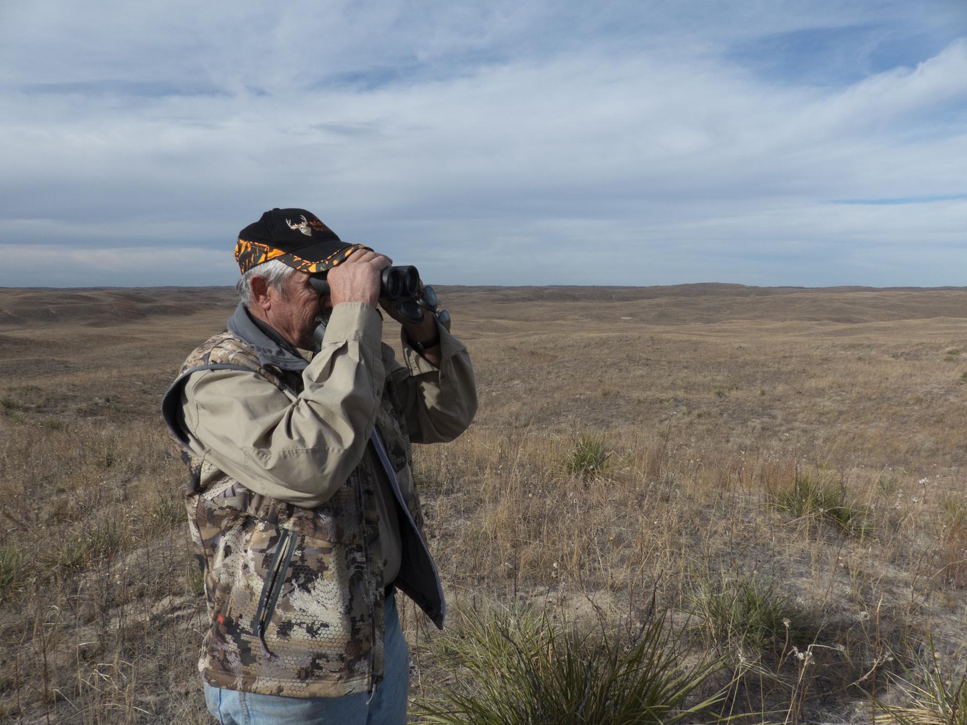 Man in camouflage gear using binoculars, scanning a vast, dry prairie under a cloudy sky.