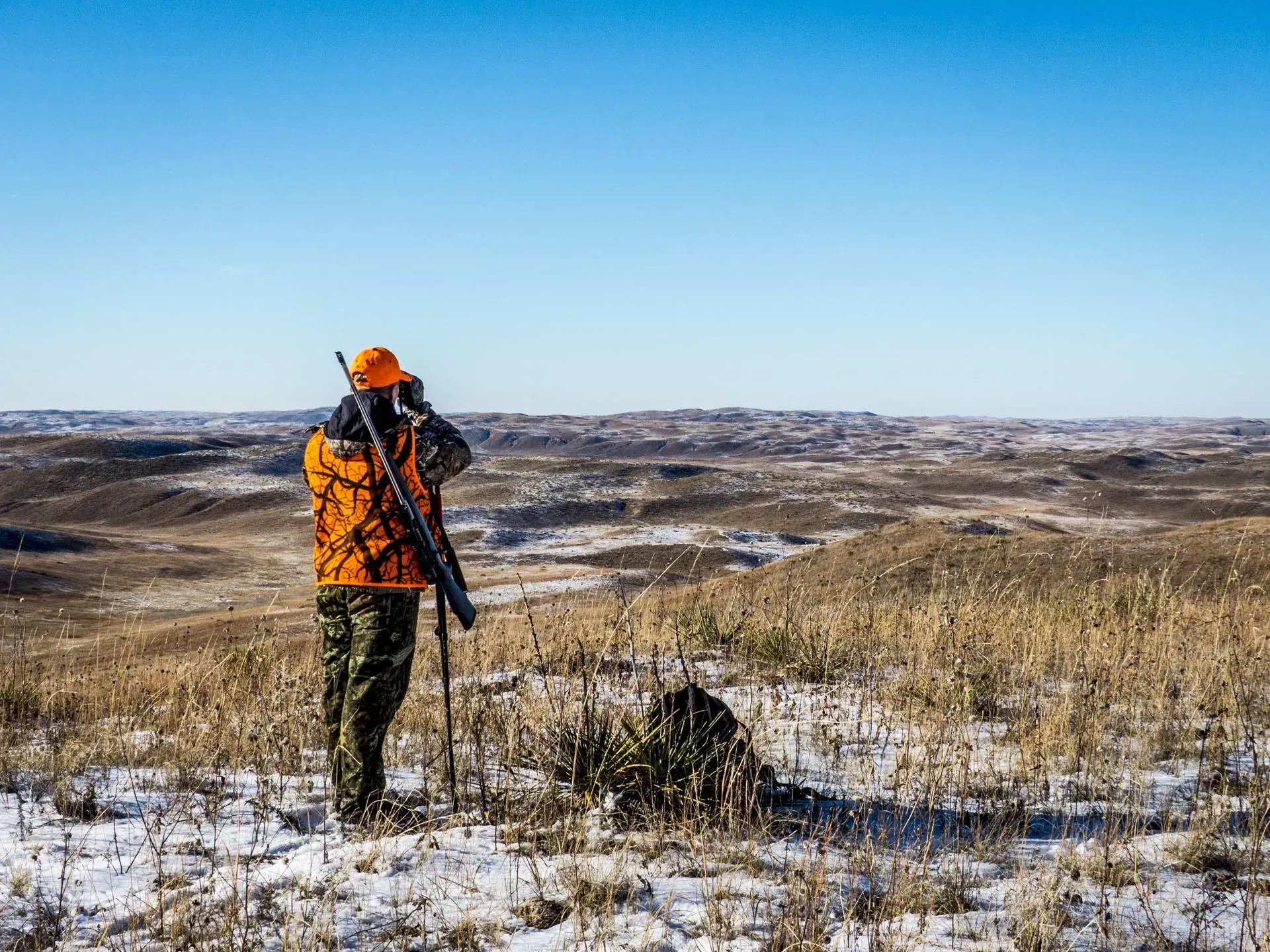 Hunter in orange vest scopes the horizon on a snowy, hilly landscape.