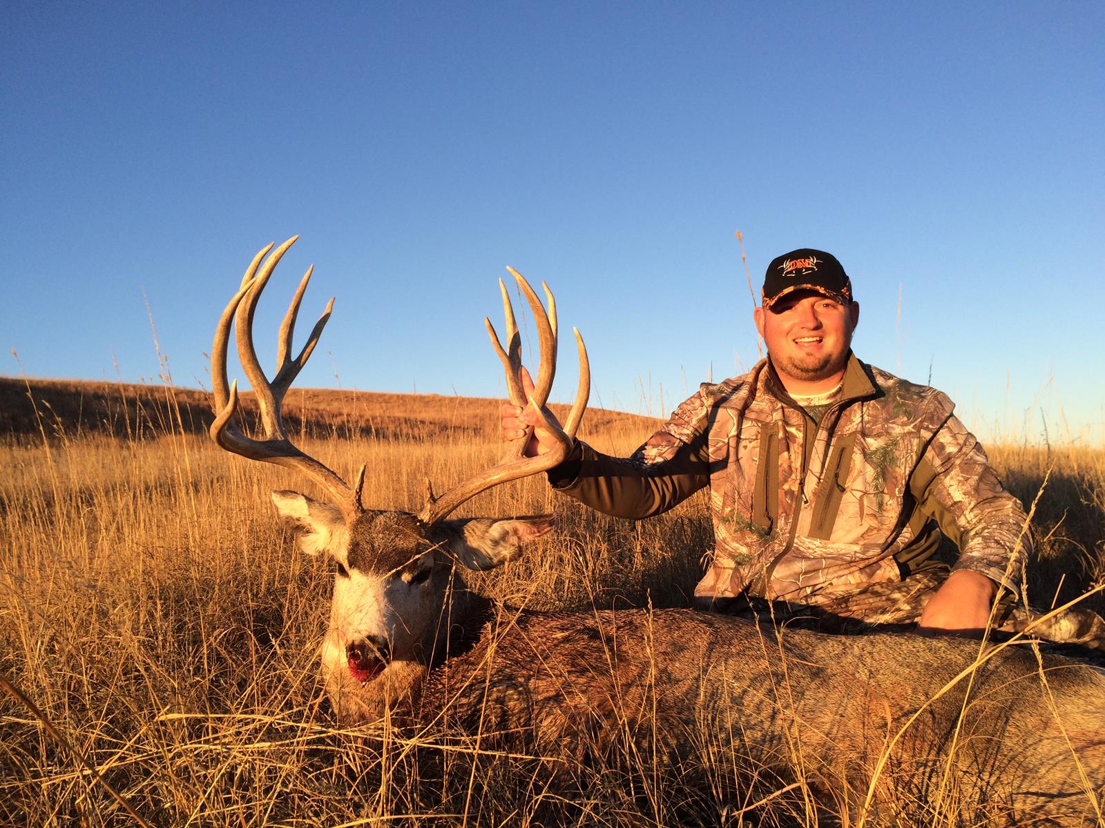 Hunter smiles next to a large buck in a field; the sky is blue and the grass is golden.