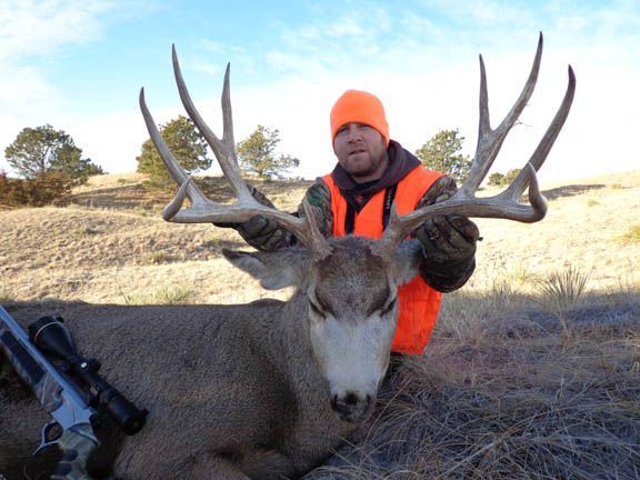 Hunter in orange vest holds large-antlered deer in a grassy field. Rifle beside the deer.