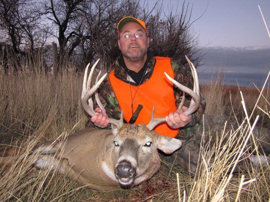 Hunter in orange vest holds a large buck with impressive antlers in a field.