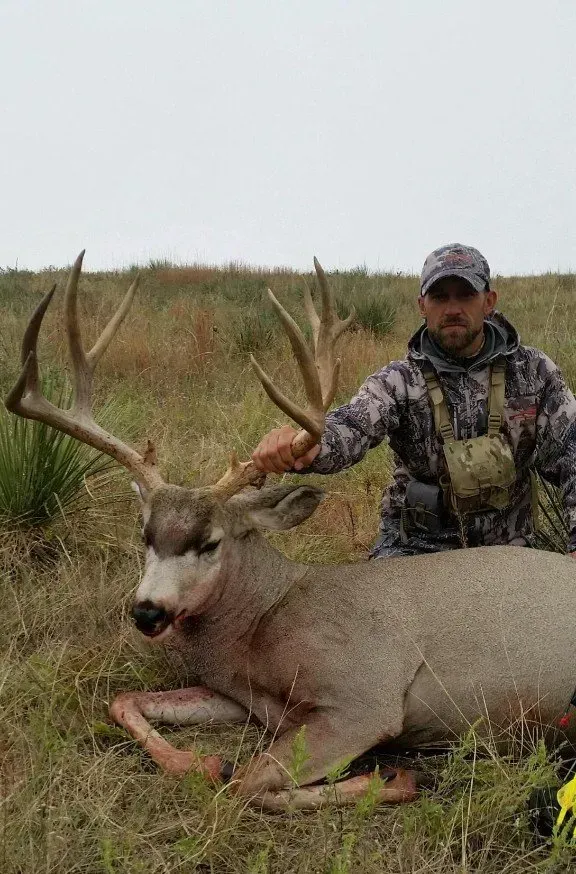 Hunter kneels beside a large buck in a field; camouflage clothing, trophy deer with large antlers.