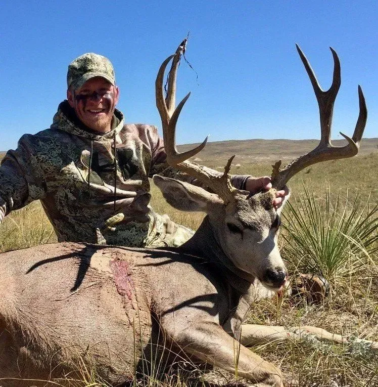 Hunter smiles, posing with a large buck in a sunny, outdoor setting.