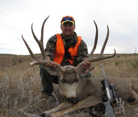 Man kneels with a large buck he hunted, wearing orange, holding rifle. Cloudy, outdoor setting.