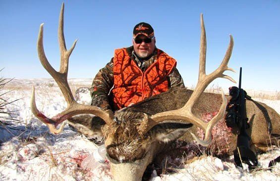 Man in orange vest poses with a large deer on snow-covered ground, rifle visible.