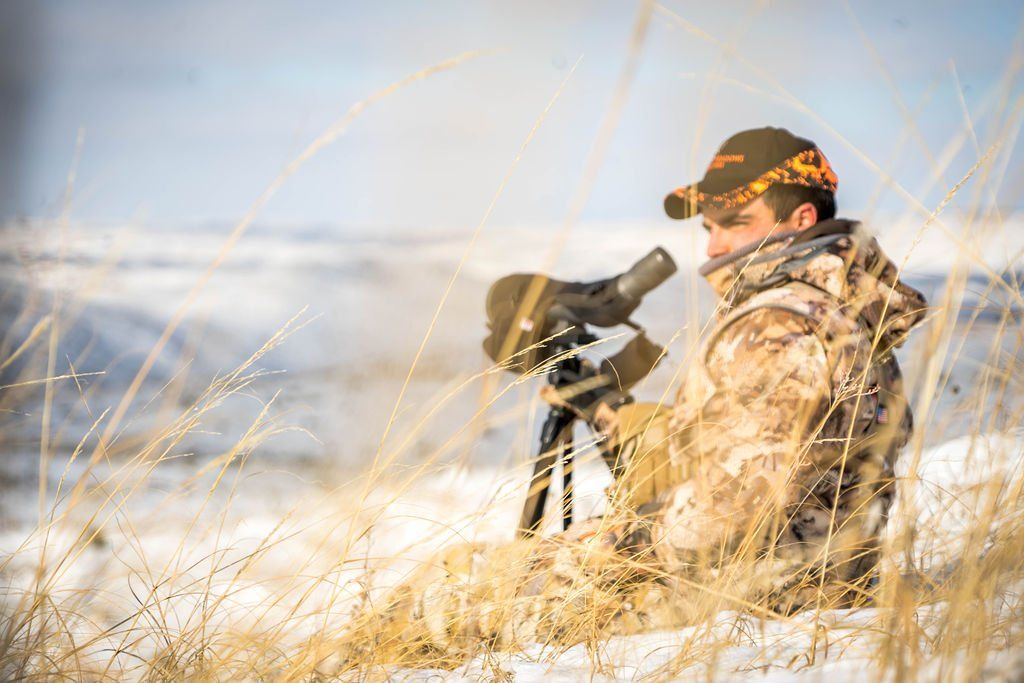 Hunter in camouflage gear with spotting scope, looking over a snowy field.