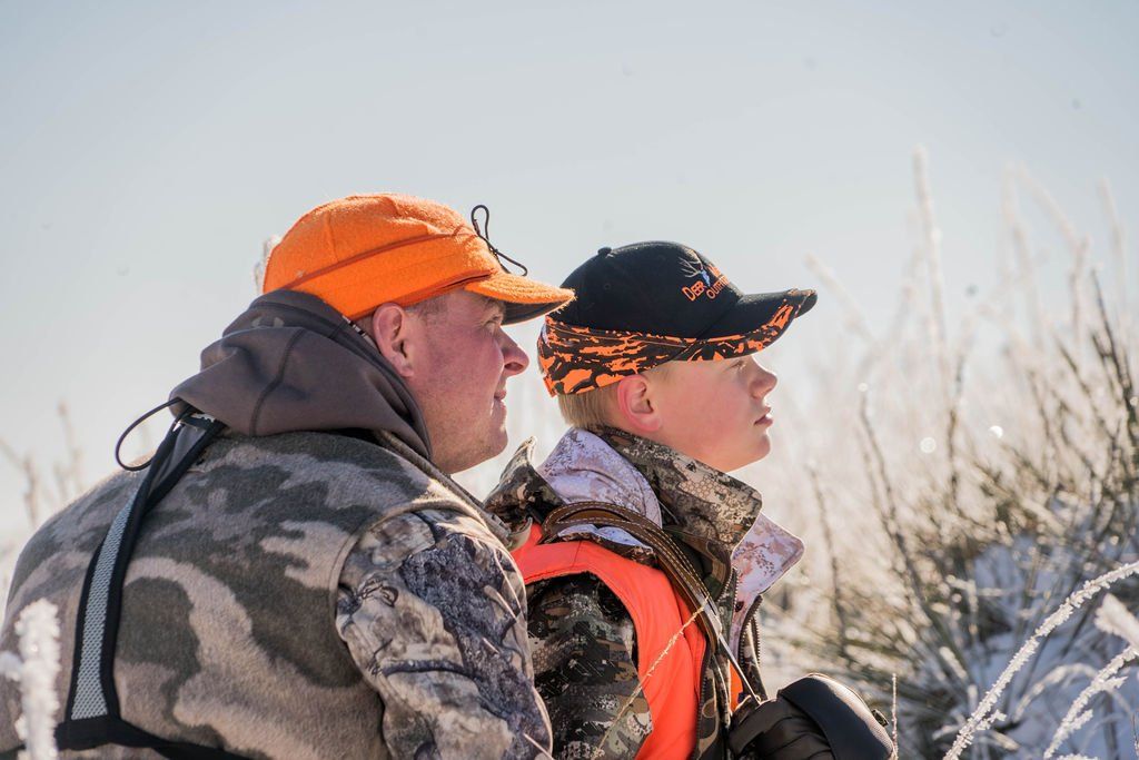 Man and youth in hunting gear looking upward, outdoors, snow, orange and camo.