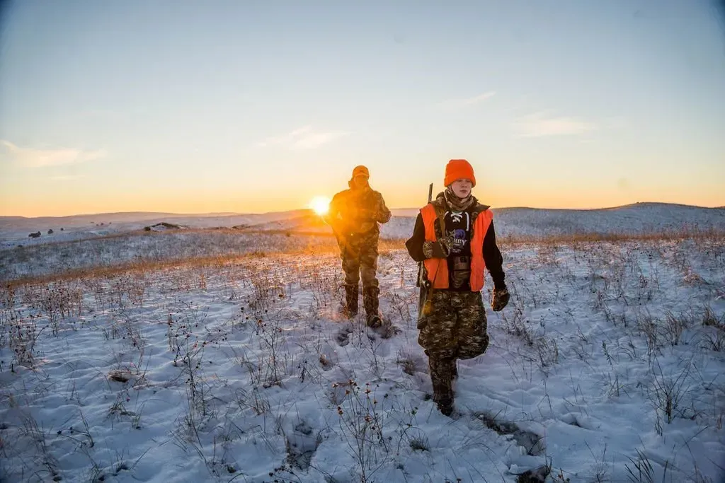 Two hunters in blaze orange and camouflage walk through snowy field at sunrise.