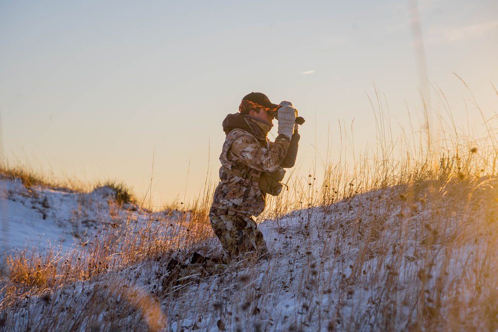 Person in camouflage kneeling in snow-covered grass, using binoculars against a golden sunset.