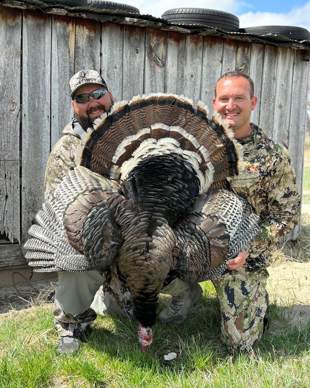Two men hold a large wild turkey, posing in front of a weathered wooden structure.