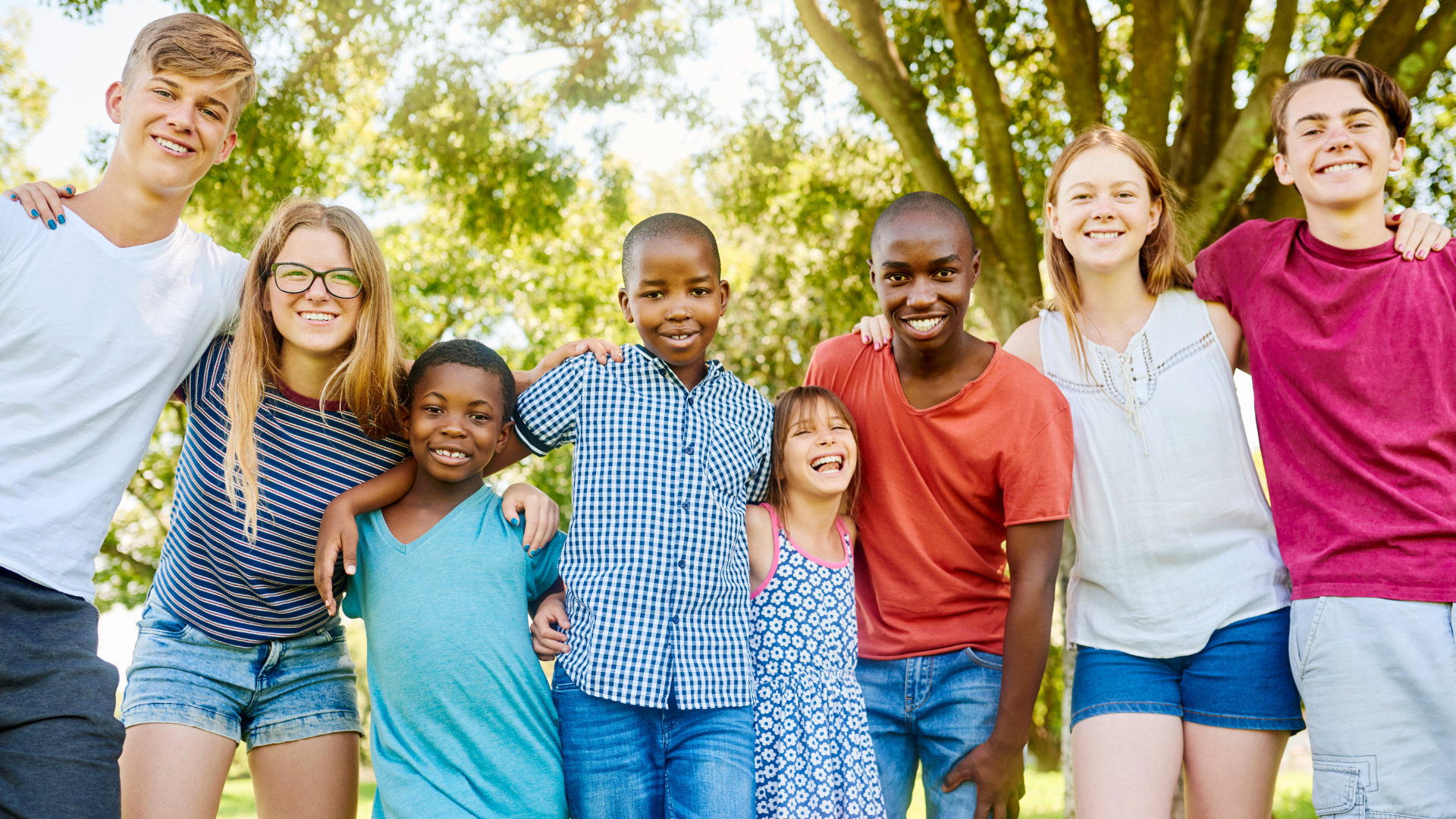 Group of people smiling, embracing outdoors under a leafy tree.