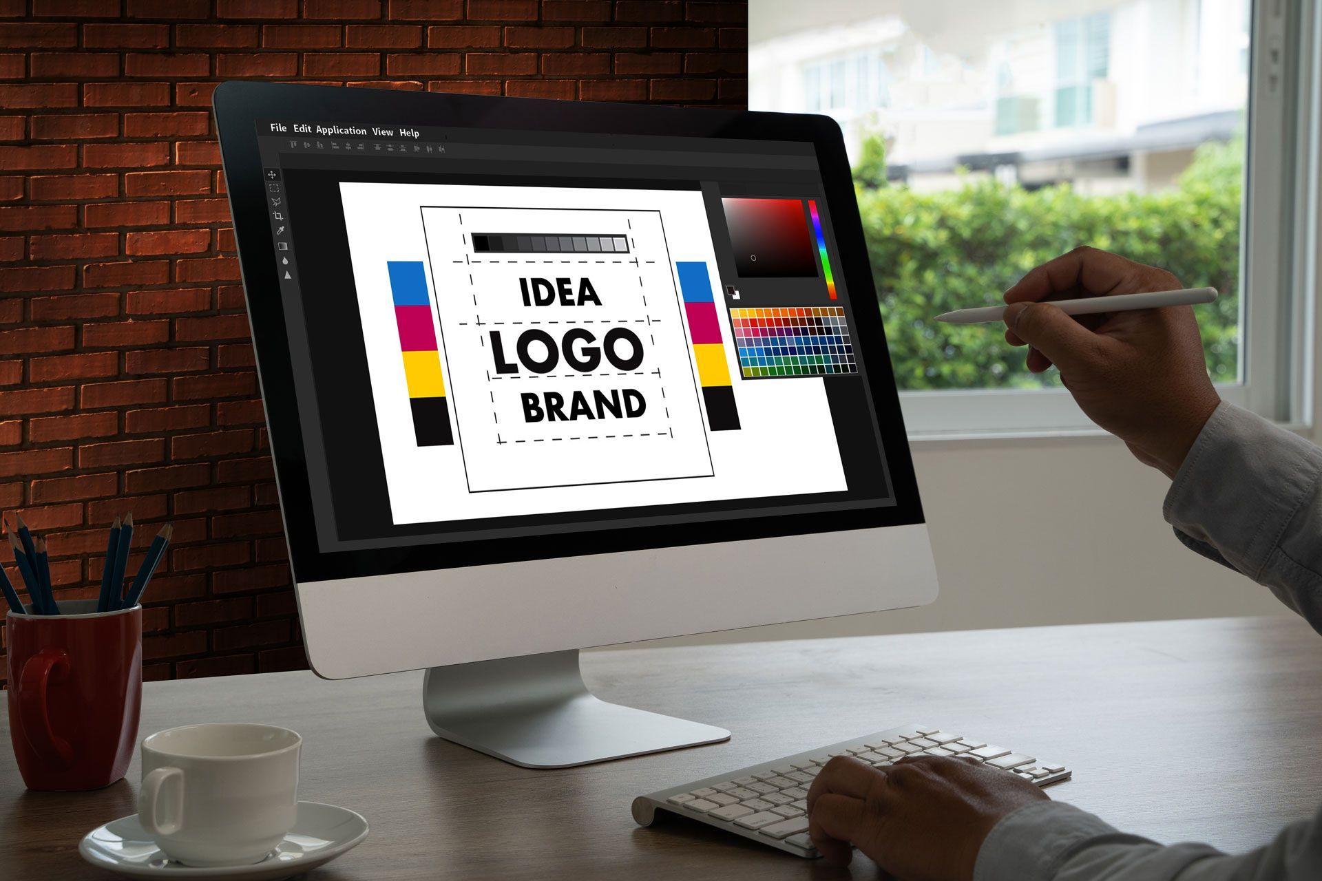 Person designing a logo on a computer at a desk. The screen displays design software with color swatches and the words “IDEA LOGO BRAND.”